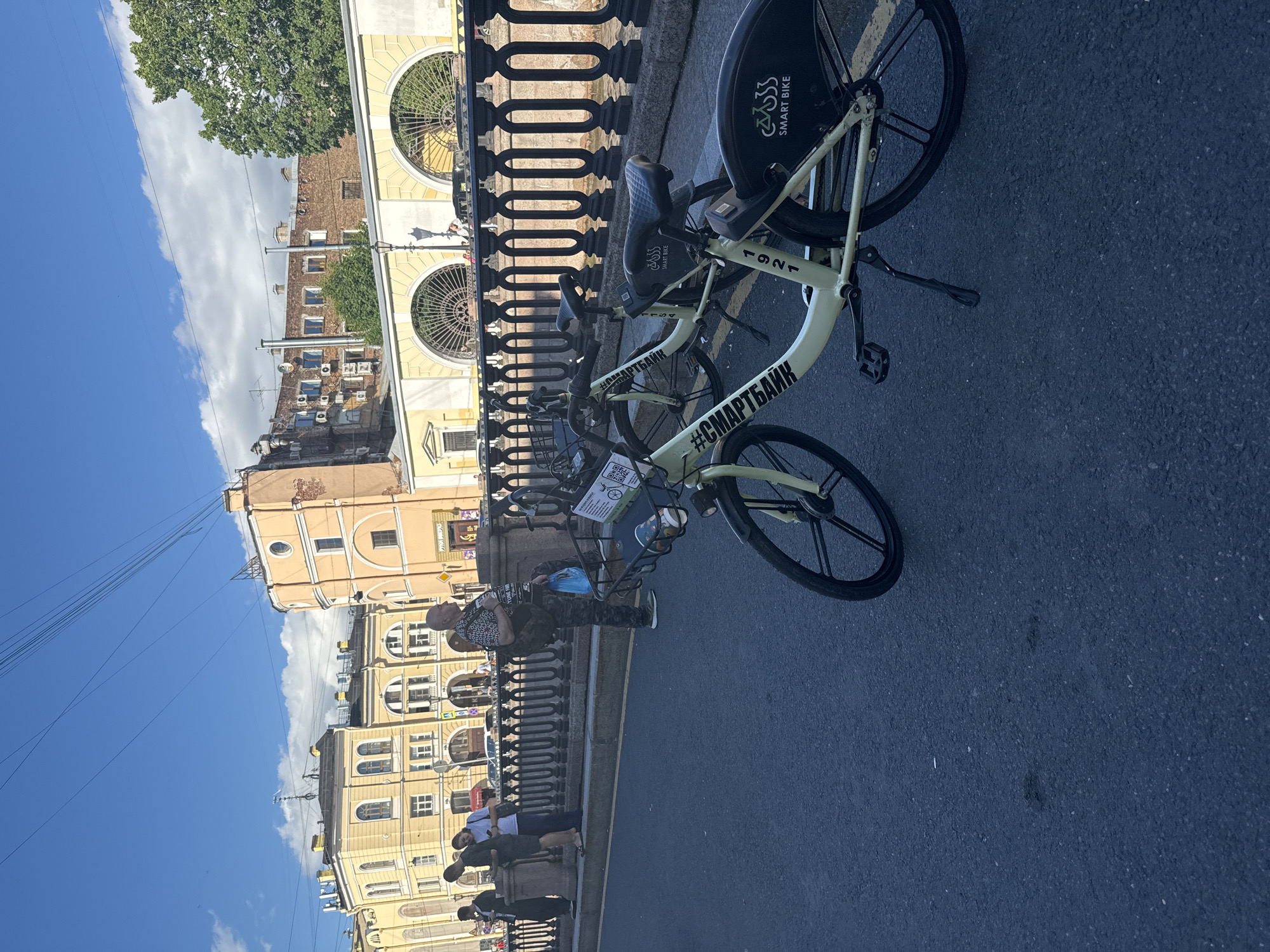 The image depicts a street scene in Saint Petersburg, Russia, likely on a bridge or embankment. In the foreground, two light-colored bicycles with "SMART BIKE" branding are parked on an asphalt road. One bicycle is more prominently featured and shows a kickstand extended. Behind the bicycles, a black metal railing runs along the edge of a raised walkway or embankment.
Several people are visible on the walkway. To the left, a group of four individuals stands near the railing, appearing to be conversing or looking at their phones. Further to the right, a man with a shaved head, wearing a patterned shirt and camouflage pants, is standing and holding a blue plastic bag. He is looking to the right.
In the background, a row of multi-story buildings lines the opposite side of the street. These buildings feature architectural details such as arched windows and ornate facades, typical of Saint Petersburg's historical architecture. The sky is blue with scattered white clouds. Power lines are visible crisscrossing the upper portion of the image. The overall impression is a daytime urban scene with elements of transportation and city infrastructure.