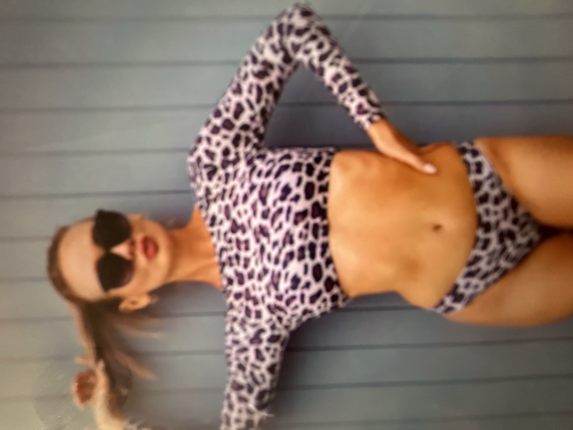 The image shows a woman lying down against a blue wooden wall, which appears to be an outdoor setting. She is wearing a leopard-print bikini, and large black sunglasses. Her right hand is resting on her hip. The lighting suggests it might be daytime. The woman's hair is pulled back. The overall impression is a relaxed, possibly summery pose. The crops provided highlight different parts of her body, the bikini's pattern, and her face with sunglasses. The background is a plain blue wall made of horizontal planks. There is no visible text in the image.