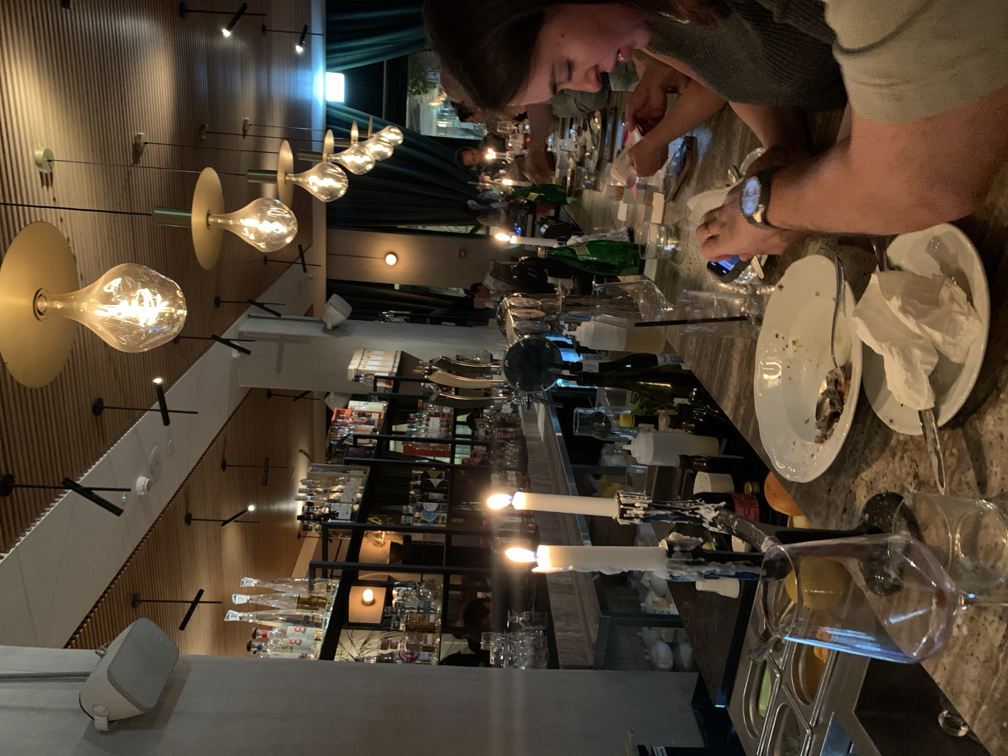 This image shows a bar scene in Tel Aviv, Israel. The focus is on a section of a bar counter where people are seated, appearing to have finished a meal. Used plates with food remnants and crumpled napkins are visible. A man's arm and wrist with a watch are near the plates, and he is holding a phone. To his left, a woman is present. Bottles of wine and glasses are scattered along the counter. In the background, a well-stocked bar shelf is visible, lined with an assortment of bottles and glassware. Candles in ornate holders burn dimly. A row of pendant light fixtures hangs from the ceiling, casting a warm glow. The room has a modern design with wood paneling and dark curtains.