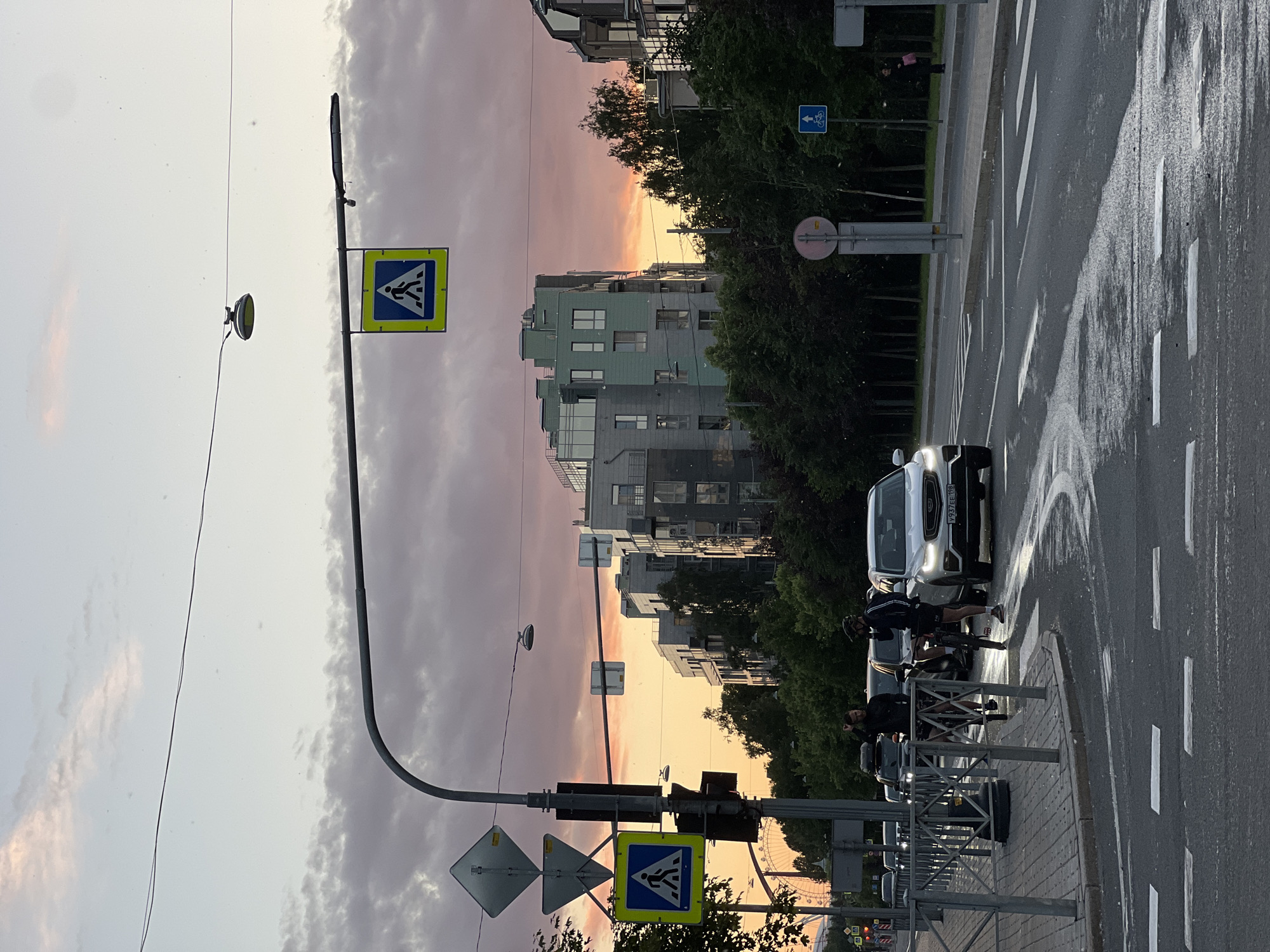 The image depicts a street scene at dusk in Saint Petersburg, Russia. The sky is painted with hues of pink and orange from the sunset, with scattered clouds. A pedestrian crossing is prominent in the foreground, with wet asphalt indicating recent rain. Two individuals on bicycles are stopped near the crossing, with one looking towards the viewer. Several cars are present on the road, including a white SUV with a license plate starting with "Y937EE 198". In the background, modern apartment buildings and a line of trees line the street. Traffic lights and pedestrian crossing signs are visible. A Ferris wheel is faintly visible in the distance on the left side of the image. The overall atmosphere is that of a city transitioning from day to night.