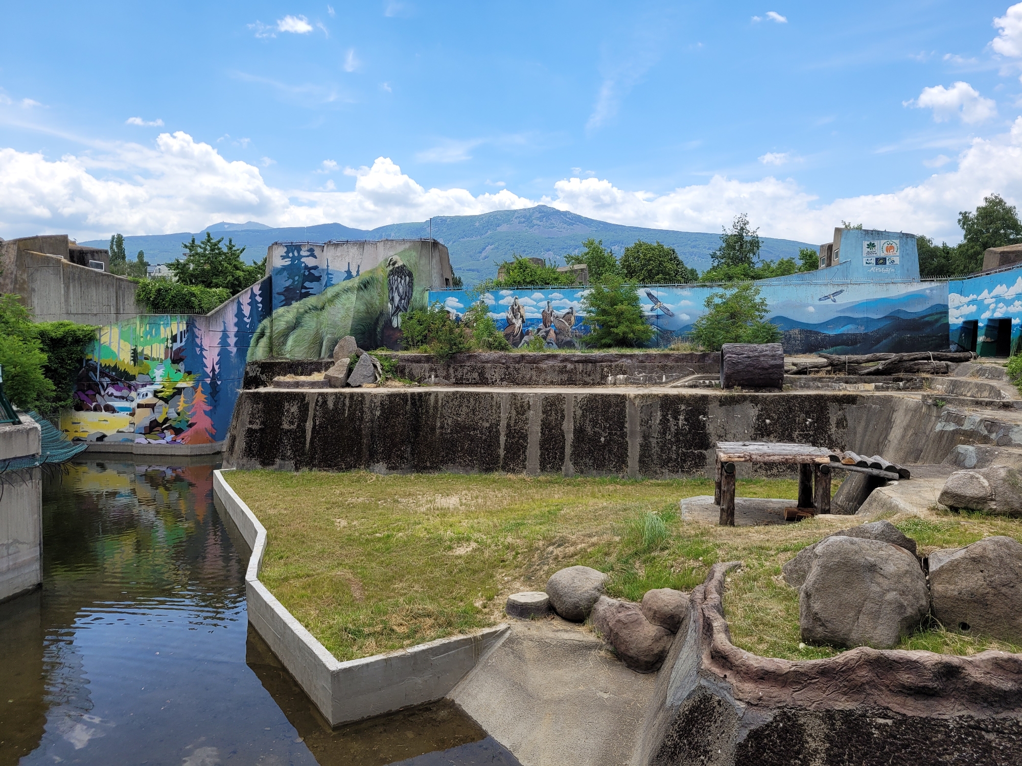 This media file shows an outdoor enclosure, likely at a zoo or wildlife park, with a large body of water in the foreground and a mountainous backdrop. The enclosure features concrete walls decorated with vibrant murals depicting wildlife and natural scenes. On the left, the mural shows a colorful, abstract landscape with trees, and further to the right, a large green dinosaur or bear is painted. In the center, a mural depicts people on horseback and birds of prey. The far right mural shows a mountainous landscape with birds flying.
The enclosure itself is designed with a mix of concrete structures, grassy areas, and large rocks, suggesting it's intended for animals that might inhabit such environments. There is a wooden structure that appears to be a shelter or feeding station for animals. The water in the foreground reflects the colorful murals and the sky.
The time of day appears to be daytime, with a bright sky filled with fluffy white clouds. The weather seems pleasant, suitable for outdoor activities. The presence of mountains in the background, along with the specific style of the murals, might offer clues to the location. The overall impression is a well-maintained and artistically decorated animal habitat. The visible text on the far right mural includes logos and what appears to be writing in Bulgarian, with the word "SOFIA" being partially legible. This strongly suggests the location is Sofia, Bulgaria, likely the Sofia Zoo. No people are visible in the scene.
