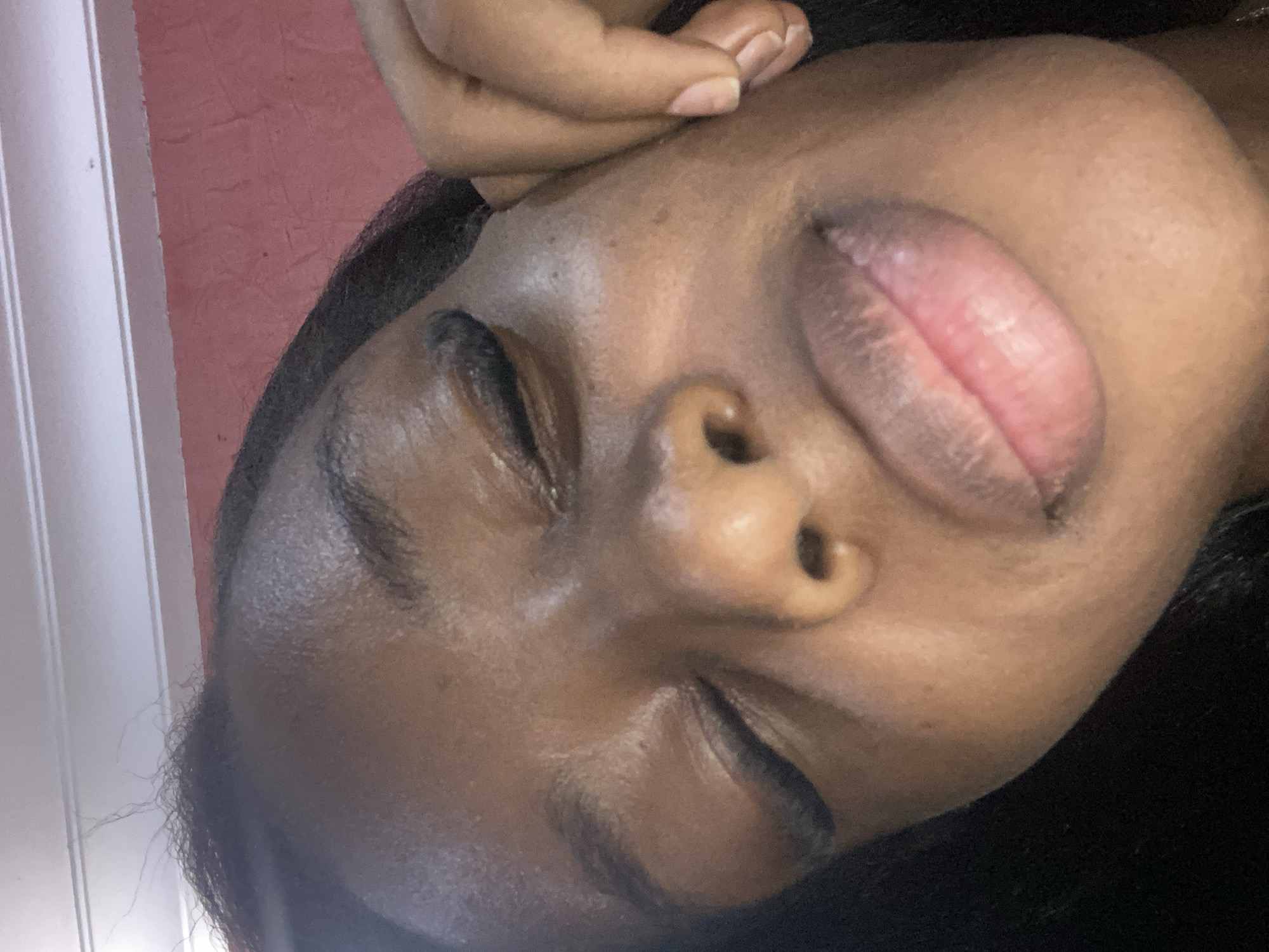 The image is a close-up, angled shot of a young Black woman's face, taken indoors. Her eyes are closed, and her head is tilted, with her right hand resting on her cheek. The background consists of a textured pink wall and a white ceiling with horizontal molding. The lighting is soft, highlighting the contours of her face and the texture of her skin. There are no discernable landmarks or clues within the image that would indicate the specific location within Jos, Nigeria.