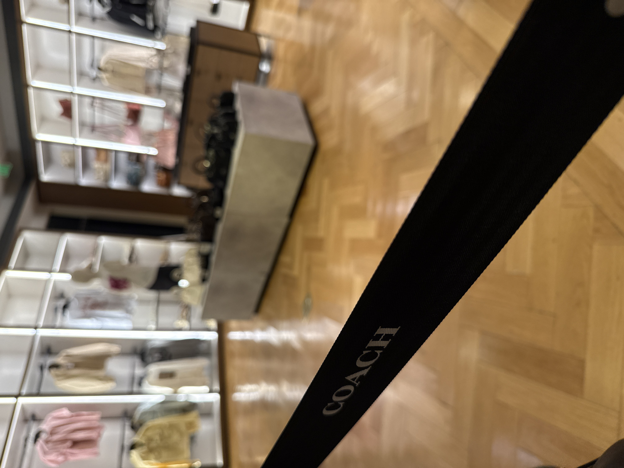 The photo depicts an overhead shot of a Coach retail store, situated in Sunrise, Florida, United States. Display shelves laden with merchandise, including bags and other accessories, are visible. A black strap with the word "COACH" printed on it is prominently in the foreground. The flooring appears to be light brown, possibly wooden, and provides a clear contrast to the black strap.