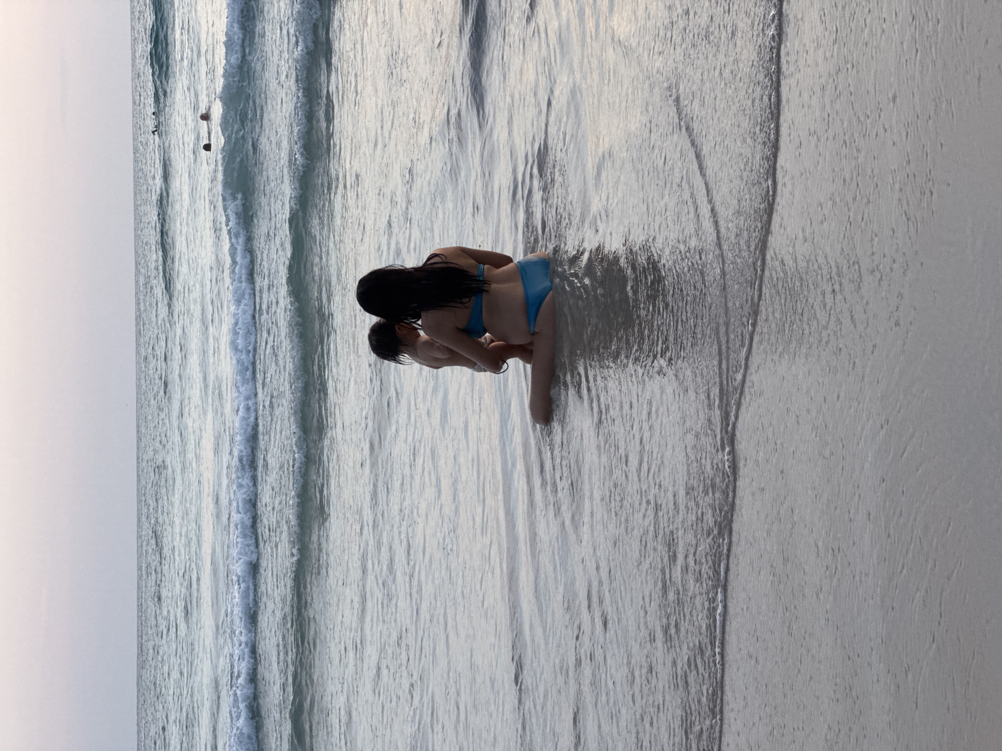 A woman in a blue bikini sits in shallow ocean water at Karon Beach, Thailand, holding a toddler. The woman has dark hair and is facing away from the camera, toward the sea. The toddler is also facing away and appears to be held in her arms. Waves gently wash around them, and the ocean extends to the horizon under a pale, hazy sky. In the distance, several other people are visible in the water, some swimming and others wading. The sandy shore is wet from the receding tide.