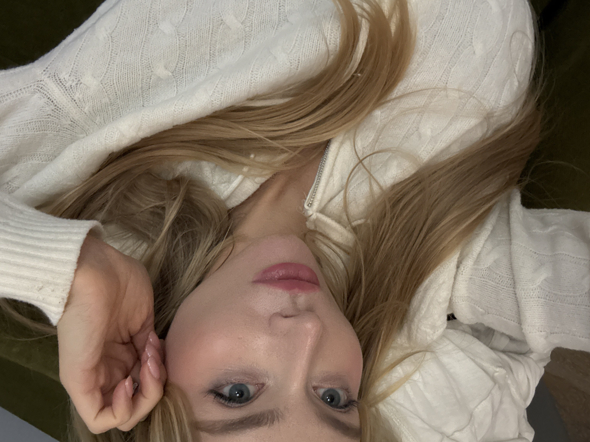 A woman with fair skin, blue eyes, and long blonde hair is lying down, wearing a white cable-knit sweater with a zipper closure. Her left hand is near her face. The background includes a green upholstered surface and a wooden floor. The scene appears to be indoors. No information suggests that the image was taken in Moscow, Russia.