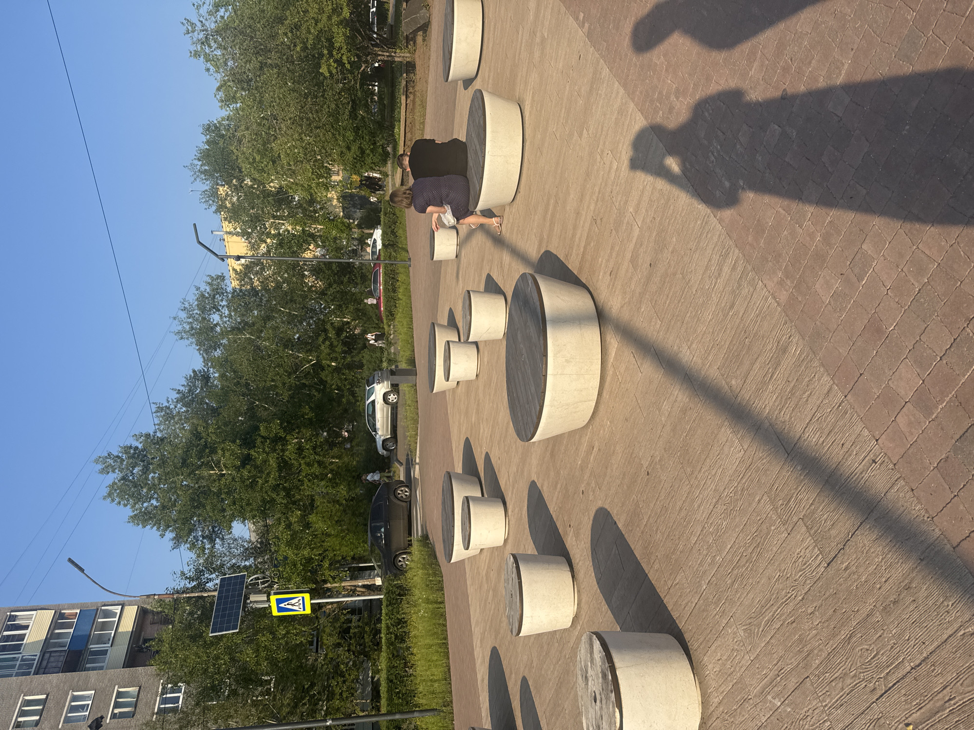The image depicts a public outdoor seating area with a paved walkway and several large, round, concrete benches with wooden tops. The benches are arranged in a circular pattern. In the background, there is a building with multiple stories and balconies, trees, a street with parked cars, and a pedestrian crossing sign with a solar panel attached to a lamppost. A couple, a man and a woman, are seated on one of the benches, facing away from the camera. Their silhouettes are cast on the ground due to the sunlight. The shadows of the person taking the photo and another individual are also visible in the foreground on the pavement. The scene appears to be in the city of Bratsk, Russia, likely during daylight hours with a clear blue sky.