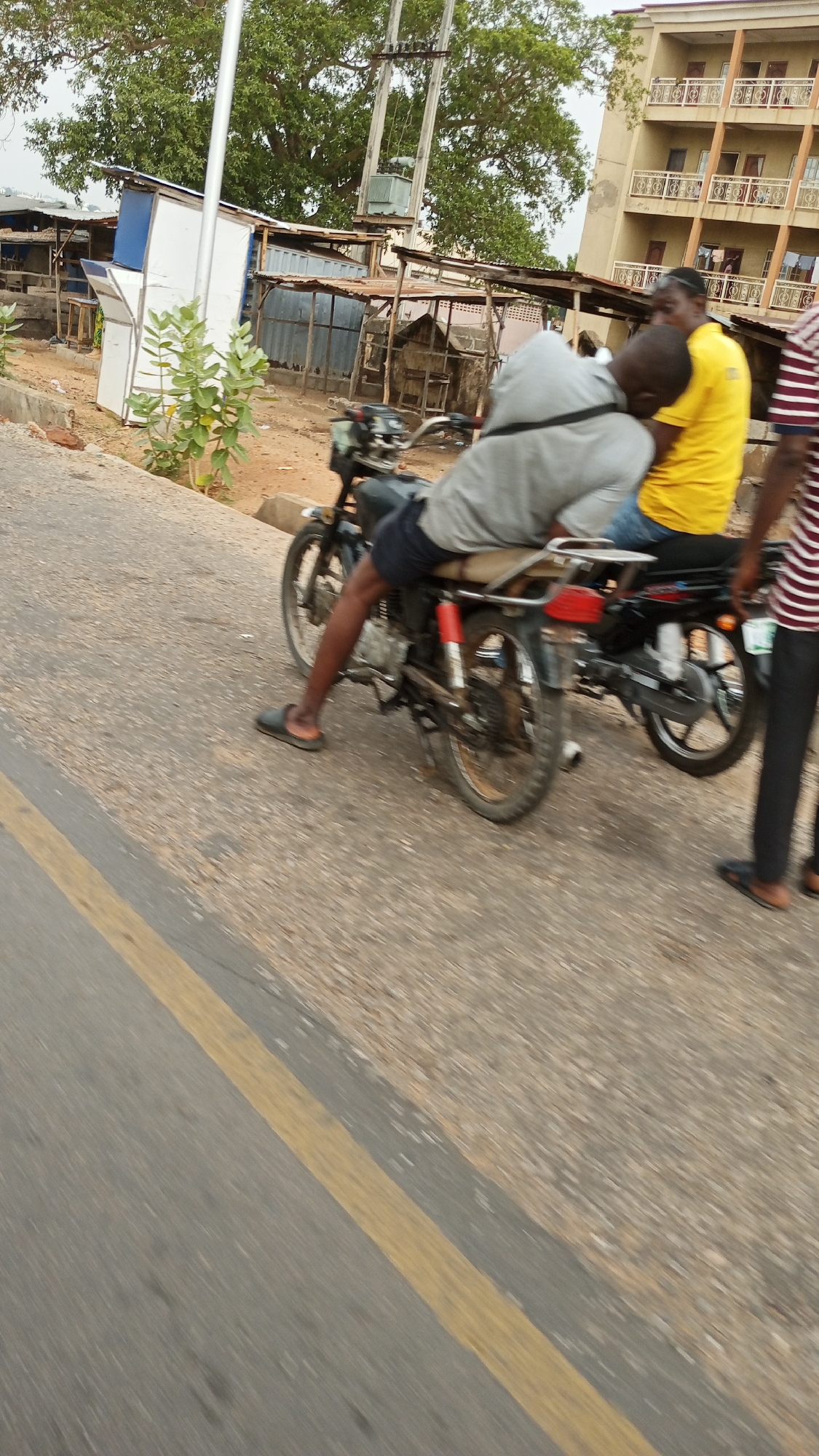 In Adankolo, Nigeria, two motorcycles are parked on a road. A person wearing a gray shirt and dark shorts sits on one motorcycle, while another person in a yellow shirt sits on the other. A third person, wearing a striped shirt and black pants, stands beside the second motorcycle. In the background, there are several small, makeshift structures and a multi-story building. The road has a yellow line marking.