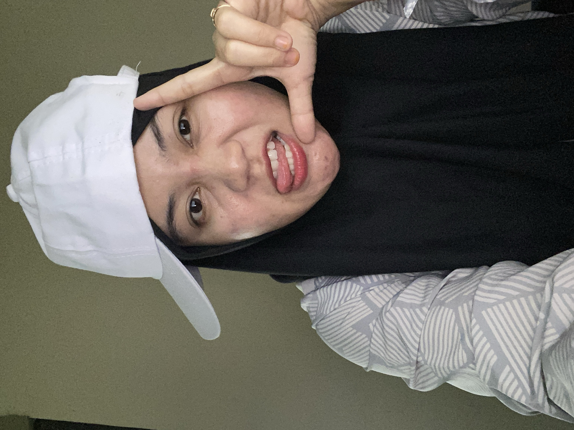 A woman is shown in a close-up, selfie-style photograph, wearing a black hijab and a white baseball cap tilted to the side. She is also wearing a patterned gray and white shirt. Her right hand is held up to her face, with her index finger and thumb forming a square shape. She is sticking her tongue out, with her mouth slightly open, revealing her teeth. A ring is visible on her right ring finger. The background is a plain, textured wall in a muted green-brown color. The lighting appears to be natural, coming from an unseen source. The overall impression is casual and playful. The image was taken in Banjarbaru, Indonesia.