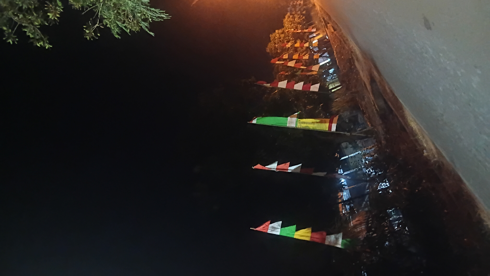 The image depicts a nighttime scene along a roadside in Prabumulih, Indonesia. A line of decorative flags, in red, white, green, and yellow, are prominently displayed along the side of a dark asphalt road. The flags are mounted on poles and appear to be triangular or pennant-shaped, suggesting a festive or celebratory occasion. Trees and foliage line the area behind the flags, with some branches from an overhanging tree visible in the upper right corner. Distant lights and reflections suggest the presence of structures or activity off-camera. The overall atmosphere is dim, with artificial lighting casting a warm glow in some areas. There are no people visible in the frame.