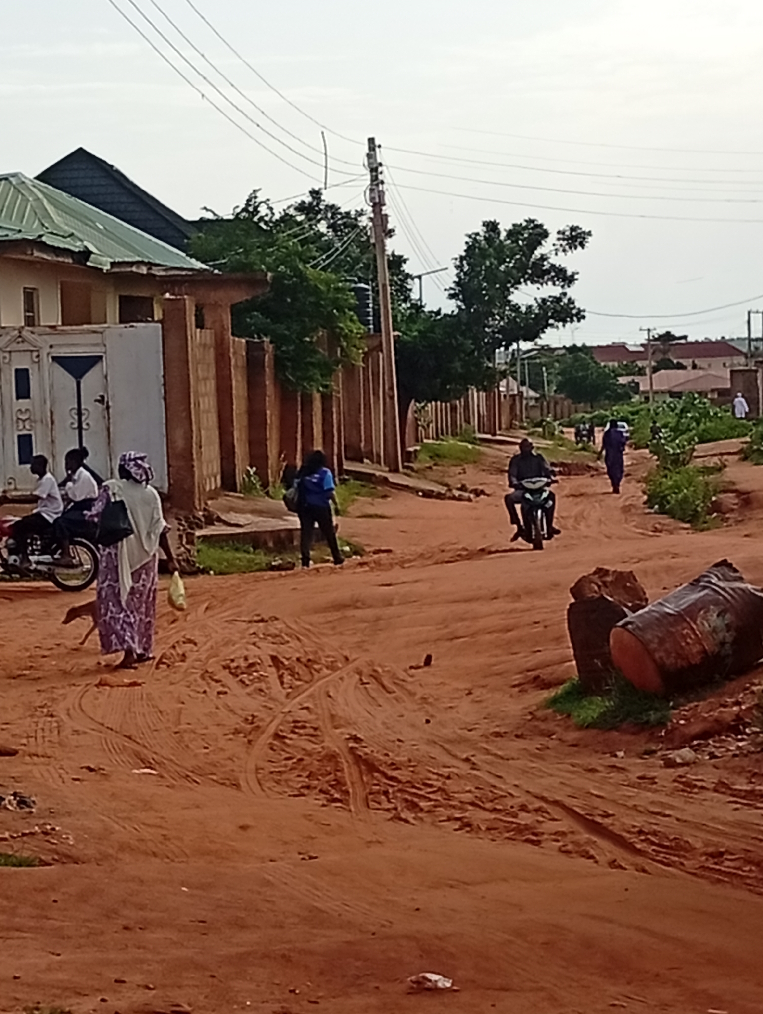 The image depicts a street scene in Tumfure, Nigeria, characterized by its unpaved, reddish-brown earth road. The sky is overcast. On the left side of the road, a fence made of reddish-brown bricks surrounds a property with a house. The house has a light-colored facade and a green, corrugated metal roof. Two young people, a boy and a girl, are seated on a motorcycle parked near a white gate. To their right, a woman wearing a patterned purple and white dress and a light-colored headscarf is walking away from the viewer, carrying a light-colored bag and a light-colored object in her right hand.
Further down the street, a person in a dark top and blue pants is walking away from the viewer, carrying a dark bag over their shoulder. On the right side of the street, a man is riding a motorcycle towards the viewer. Another person in a dark blue garment is walking away from the viewer in the distance. The road is rutted, indicating recent vehicle traffic. In the foreground, two large, rusted metal barrels lie on the side of the road, partially covered by vegetation. The environment appears to be a residential area with houses and trees visible along the street.