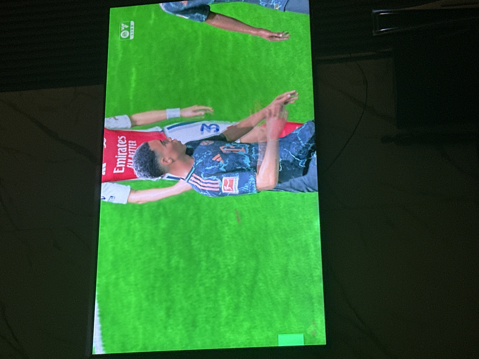 This is a media file depicting a video game scene, likely a soccer match. The screen displays a bright green field, typical of a soccer pitch. Two individuals, appearing to be soccer players rendered in a video game style, are the central focus. One player is lying on the ground, with their arms outstretched. The other player is bent over, possibly reaching out to the player on the ground. Both players are wearing soccer uniforms. The player on the ground is wearing a blue jersey with intricate patterns and the number "11" visible. The other player is partially visible, wearing a red jersey with the "Emirates FLY BETTER" logo. There are also some graphical elements present on the screen, including a small green rectangle in the bottom right corner, and a logo with the number "7" and the word "LIVE" in the top left corner, which is commonly seen in sports broadcasts or video games. The overall impression is a still frame from a soccer video game, capturing a moment of interaction between two players on the field. The lighting appears to be bright, suggesting an indoor setting or a well-lit screen. The image is taken from an angle, showing the edge of the screen.