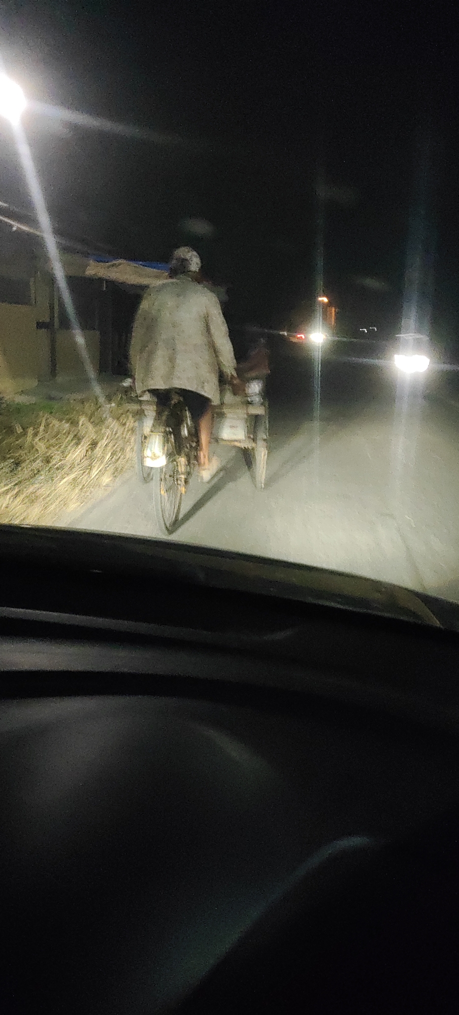 An individual is riding a three-wheeled bicycle, often referred to as a becak, on a paved road at night in Manyar, Indonesia. The becak is pulling a cart behind it. The person on the becak is wearing a light-colored jacket and appears to have a hat on. The scene is illuminated by streetlights and the headlights of a vehicle, likely the one from which the photo was taken. There are lights and some structures visible in the background, suggesting a semi-urban or rural setting. The road surface appears to be asphalt, and there is dry vegetation along the side of the road.