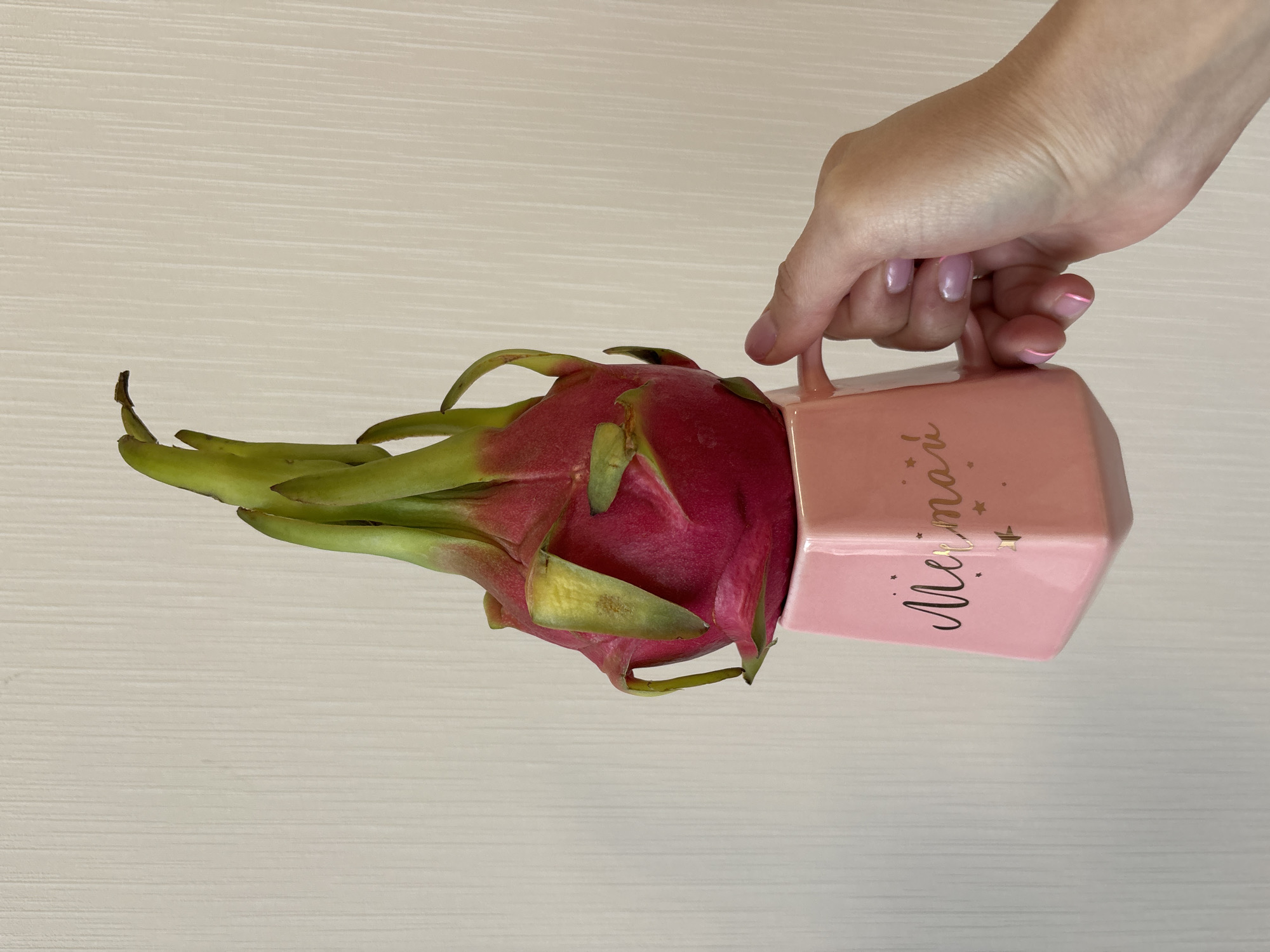 A horizontal, top-down shot shows a hand holding a light pink, square-shaped mug filled with a pink dragon fruit on a light-colored, horizontal surface. The dragon fruit is positioned with its pink flesh inside the mug and green protrusions extending outward. The mug has a handle on one side and the word "Mermaid" written in gold cursive on its side. A hand with pink-tipped fingernails holds the mug by its handle. The background is a light surface with faint horizontal graining. This image was taken indoors, at an unknown location. The city of Izhevsk, Russia is not visible in this image.