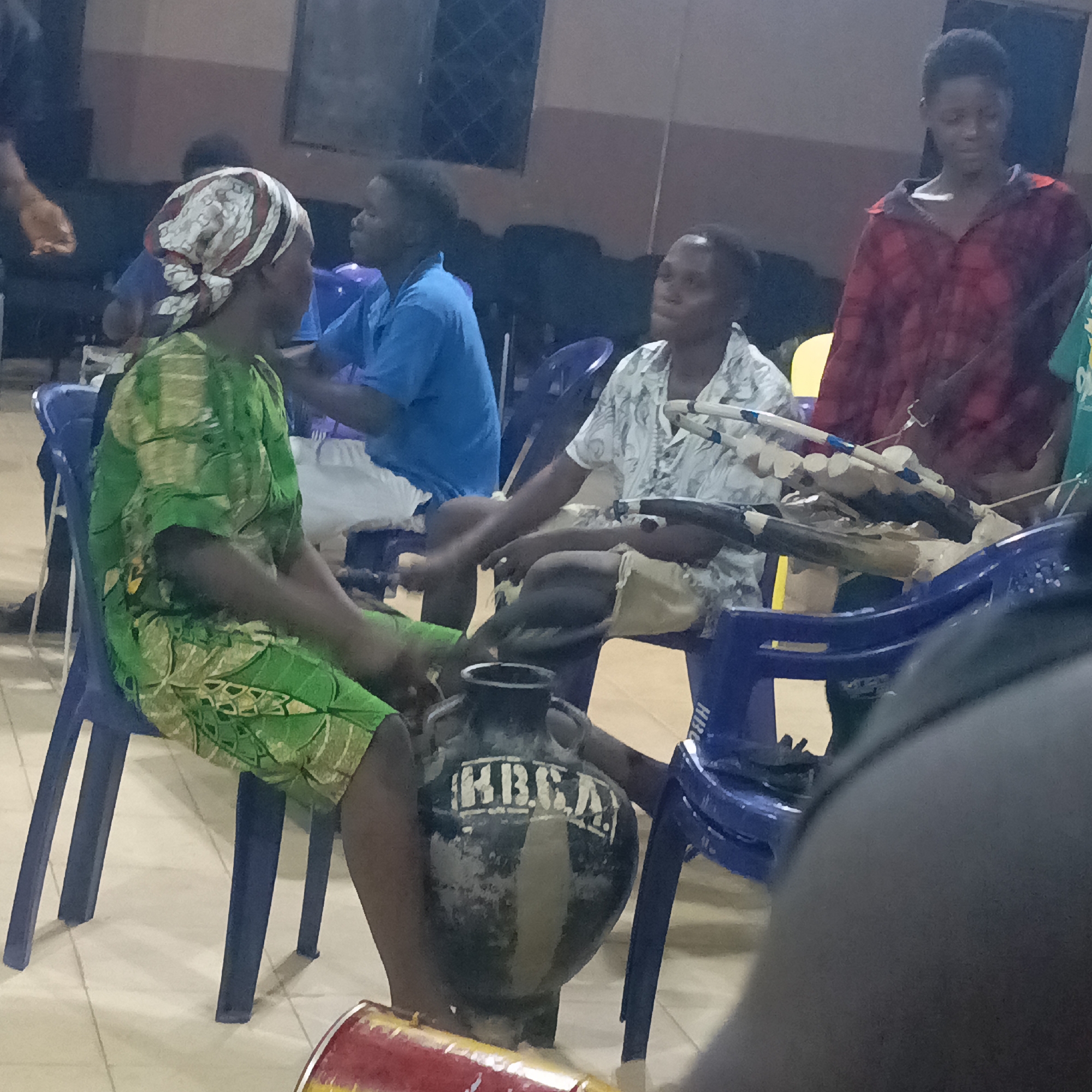 The image shows a group of people indoors, likely in a public space in Akwanga, Nigeria. A woman wearing a green patterned dress and a headscarf sits on a blue plastic chair. Next to her, another person sits with a string instrument resting on their lap. A large, decorative vase with the inscription "HBCA" stands between them. Other individuals are visible in the background, some seated and one standing in a red plaid shirt. The room appears to be dimly lit with tiled flooring.