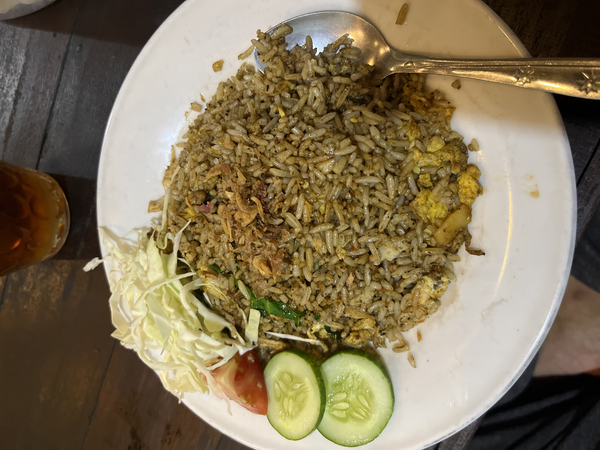 A close-up, overhead shot displays a plate of nasi goreng, a popular Indonesian fried rice dish, on a dark wooden table. The rice is a deep brown color, mixed with visible pieces of egg and what appear to be small bits of meat or vegetables. A garnish of thinly sliced fried onions sits atop the rice. To the left of the plate are three garnishes: shredded white cabbage, a slice of red tomato, and two circular slices of green cucumber. A silver spoon rests on the right side of the plate, partially submerged in the rice. In the background, a glass of dark amber liquid, likely iced tea, is visible. The overall scene suggests a casual dining setting in Tampingan, Indonesia.