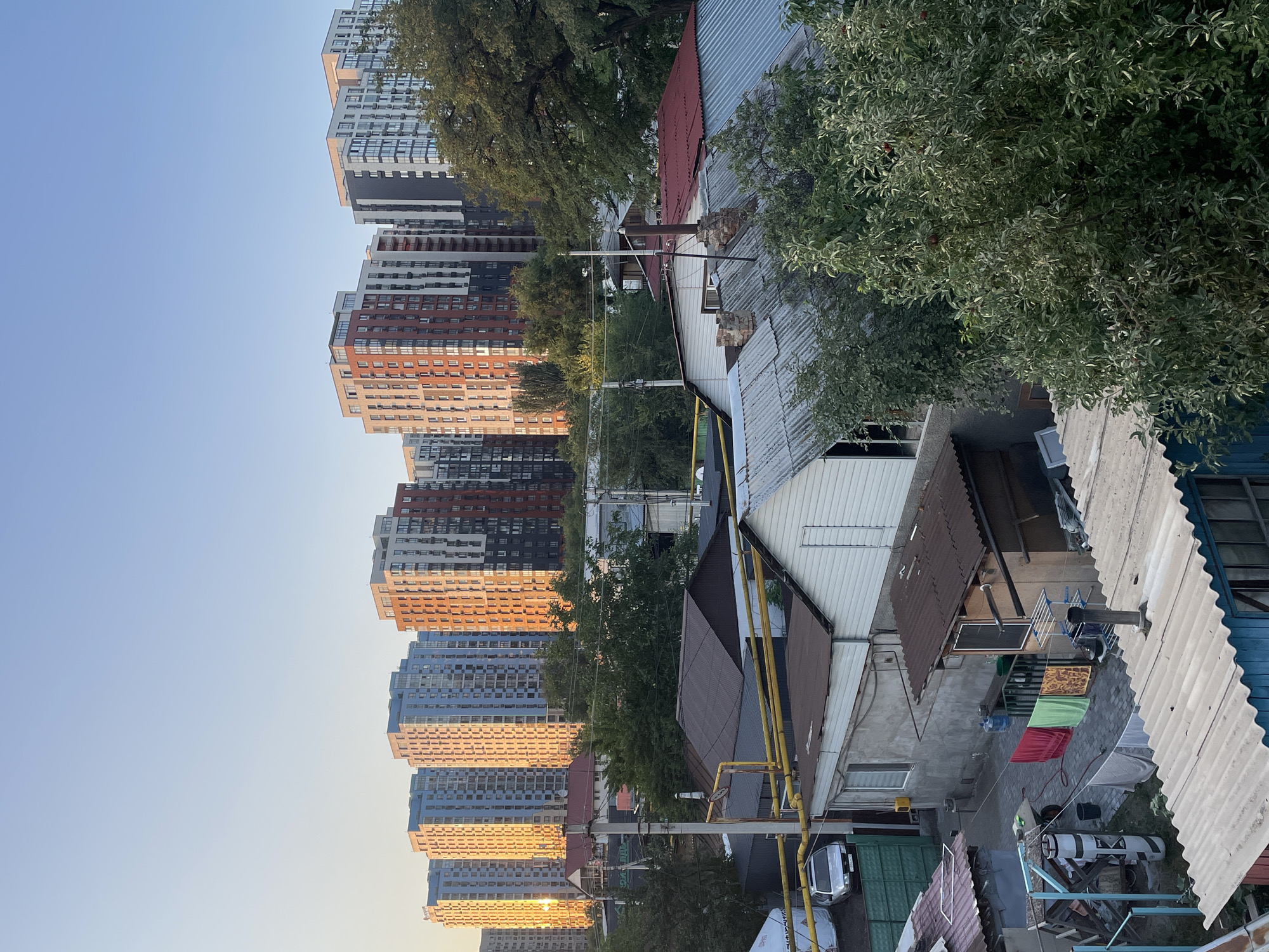 The image depicts a view of a residential area in Almaty, Kazakhstan, with modern high-rise apartment buildings in the background casting long shadows across a foreground of older, single-story structures. The time of day appears to be late afternoon or early evening, as indicated by the warm golden light illuminating the facades of the distant buildings.
In the foreground, a cluster of low-rise buildings with corrugated metal roofs are visible. These structures appear to be older and more utilitarian, some showing signs of wear and tear. Clothes are seen hanging on a line between two buildings, suggesting active residential use. A silver car is parked next to a green metal fence in the left portion of the scene.
Behind these older buildings, a line of modern apartment blocks stretches across the horizon. These buildings are varied in design, with some featuring light blue facades and others a mix of red, gray, and white. The sunlight reflects brightly off the windows and walls of these taller structures. Trees are interspersed throughout the scene, with dense green foliage creating a contrast with the urban architecture. The sky is clear and a pale blue.
