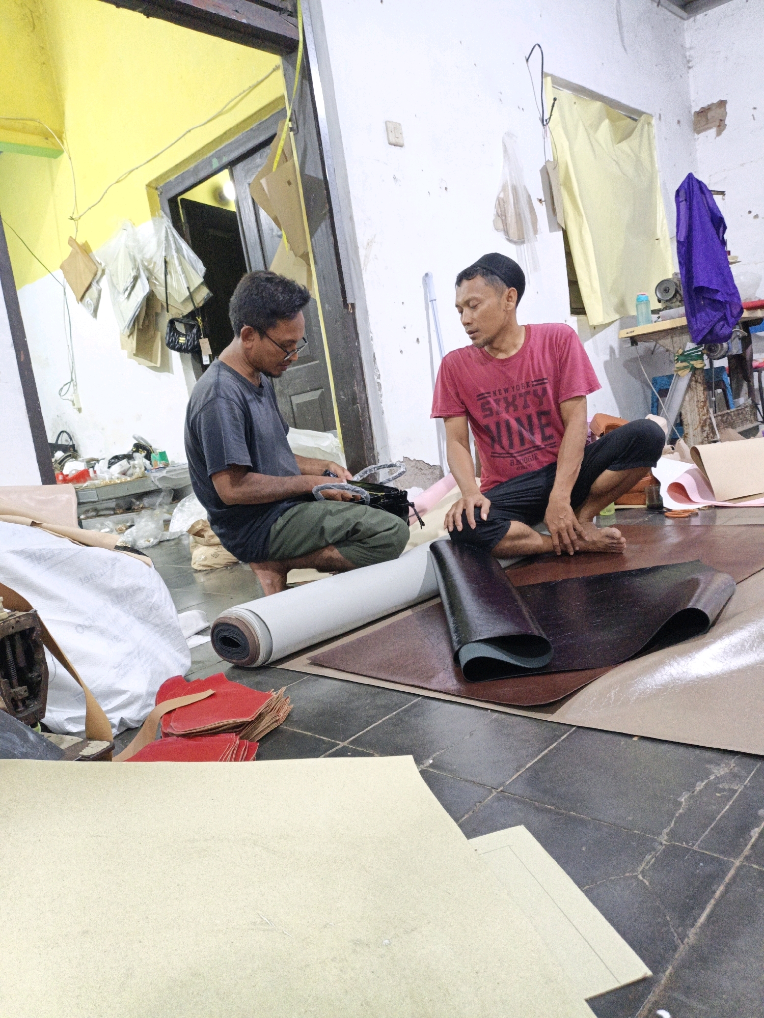 Two men are in a workshop in Mangli, Indonesia. The man on the left, wearing a dark t-shirt and green pants, is looking down at something in his hands. He has glasses and short, dark hair. The man on the right, wearing a red t-shirt and black pants, is sitting cross-legged and looking towards the man on the left. He is wearing a black cap.
The floor is tiled and scattered with various materials, including large rolls of what appears to be leather or vinyl, sheets of paper, and red fabric pieces. There is a sewing machine visible in the background to the right, along with tools and supplies. The walls are white, and the ceiling is painted yellow. The scene suggests a workspace for crafting or manufacturing leather goods.