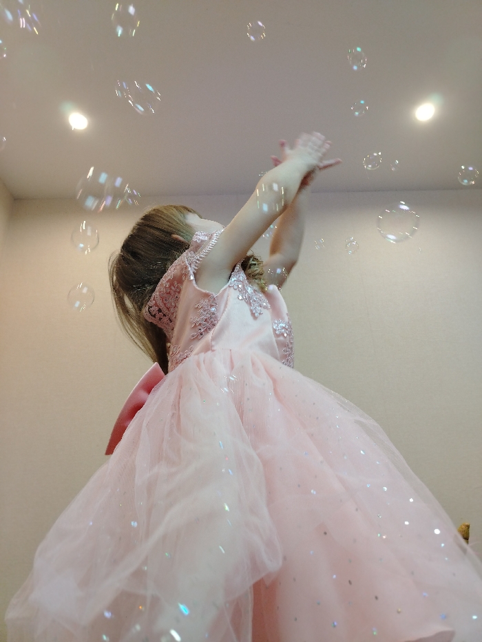 The image captures a low-angle view of a young person, likely a girl, joyfully interacting with bubbles indoors. The subject is seen from the side, looking upwards and reaching with both hands towards numerous clear, iridescent bubbles floating in the air.
She is wearing an elaborate, light pink dress, possibly a gown. The bodice of the dress appears to have delicate lace or embroidered details on the shoulders and upper chest, with short, cap-like sleeves. The skirt is voluminous, made of multiple layers of soft, sheer pink tulle or netting, and is embellished with small, shimmering sparkles that catch the light, adding to a whimsical effect. A large pink bow or sash is partially visible at the back of her waist. Her hair, which appears to be long and light brown or auburn, flows down her back.
The setting is indoors, characterized by plain, light-colored walls and a flat ceiling. Two recessed ceiling lights are visible, with one creating a soft glow or lens flare in the upper left part of the image. The lighting appears to be artificial and diffused, highlighting the bubbles' reflective surfaces.
The overall impression is one of playful wonder and excitement, as the child stretches out to touch or catch the ethereal bubbles that fill the upper part of the frame. No text is visible in the image.
