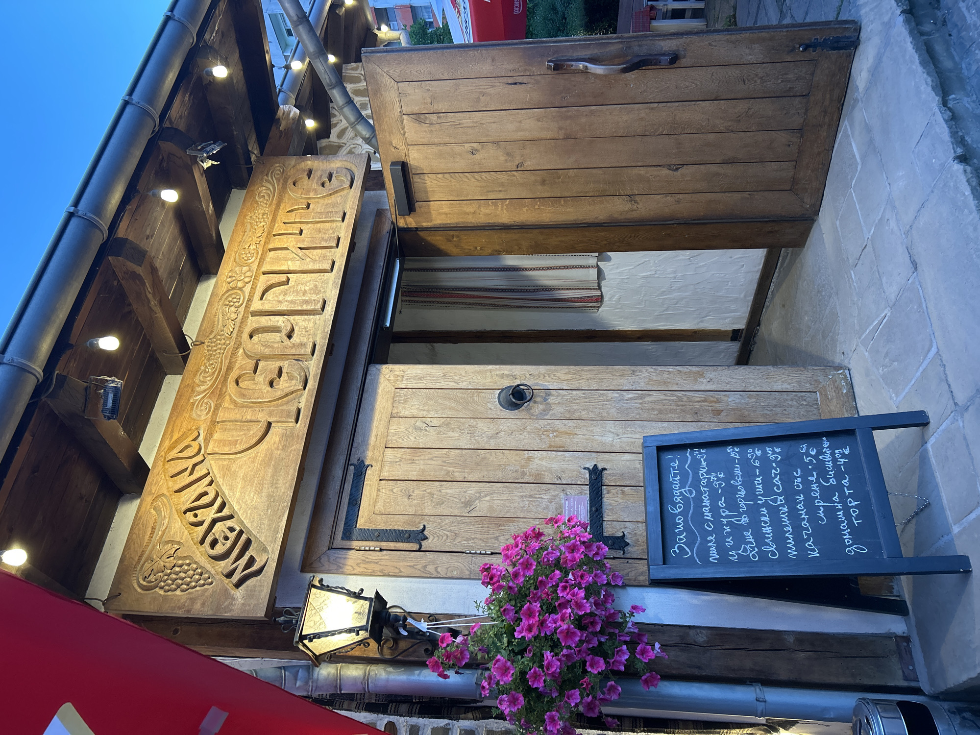 The image depicts the entrance to a traditional Bulgarian restaurant, likely located in Sofia, Bulgaria, given the Cyrillic signage. The facade features rustic wooden doors, one slightly ajar, revealing a glimpse of an interior adorned with a traditional woven fabric. Above the entrance, a large, intricately carved wooden sign displays the restaurant's name in Cyrillic. A wrought-iron lantern hangs to the left, casting a warm glow, and a pot of vibrant pink flowers adds a touch of color to the scene. A black A-frame chalkboard sign stands on the cobblestone pavement, displaying a menu written in chalk, also in Cyrillic. The overall ambiance is cozy and inviting, with strings of light bulbs illuminating the wooden beams of the building's overhang. The setting suggests an evening time, with the sky a deep blue, indicating dusk or early night.