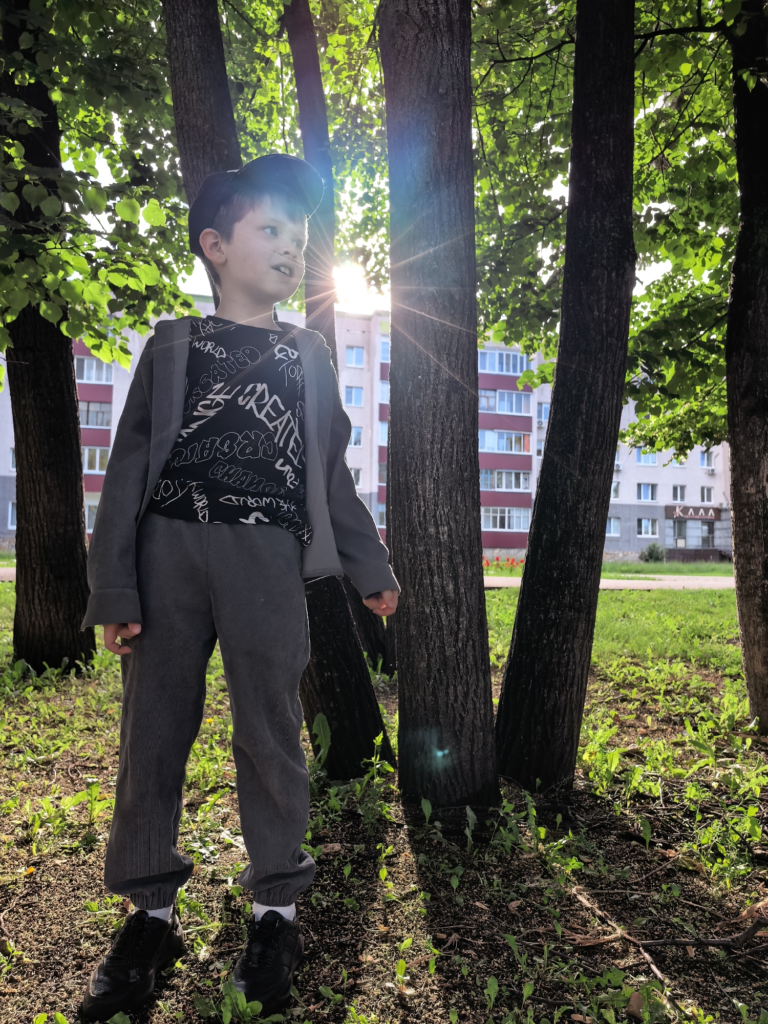 A young boy stands in a park-like setting, surrounded by several trees. He is wearing a black baseball cap, a grey jacket over a black t-shirt with white text, and grey pants. His black sneakers are visible. The boy is looking to his left with a slight smile. Sunlight streams through the leaves of the trees, creating dappled light and shadows on the ground and on the boy. In the background, beyond the trees, a multi-story apartment building with balconies and windows is visible. The ground is covered with grass, fallen leaves, and small plants. The scene suggests it is daytime, likely late afternoon or early evening, given the low angle of the sun and the warm light. The location cues, such as the apartment building style and the vegetation, along with the provided location context of Sterlitamak, Russia, help place the scene. The boy's expression conveys a sense of wonder or engagement with something off-camera. The text on the boy's t-shirt appears to be in English and includes words like "WORLD" and "CREATE," though it is partially obscured and difficult to read in full.
