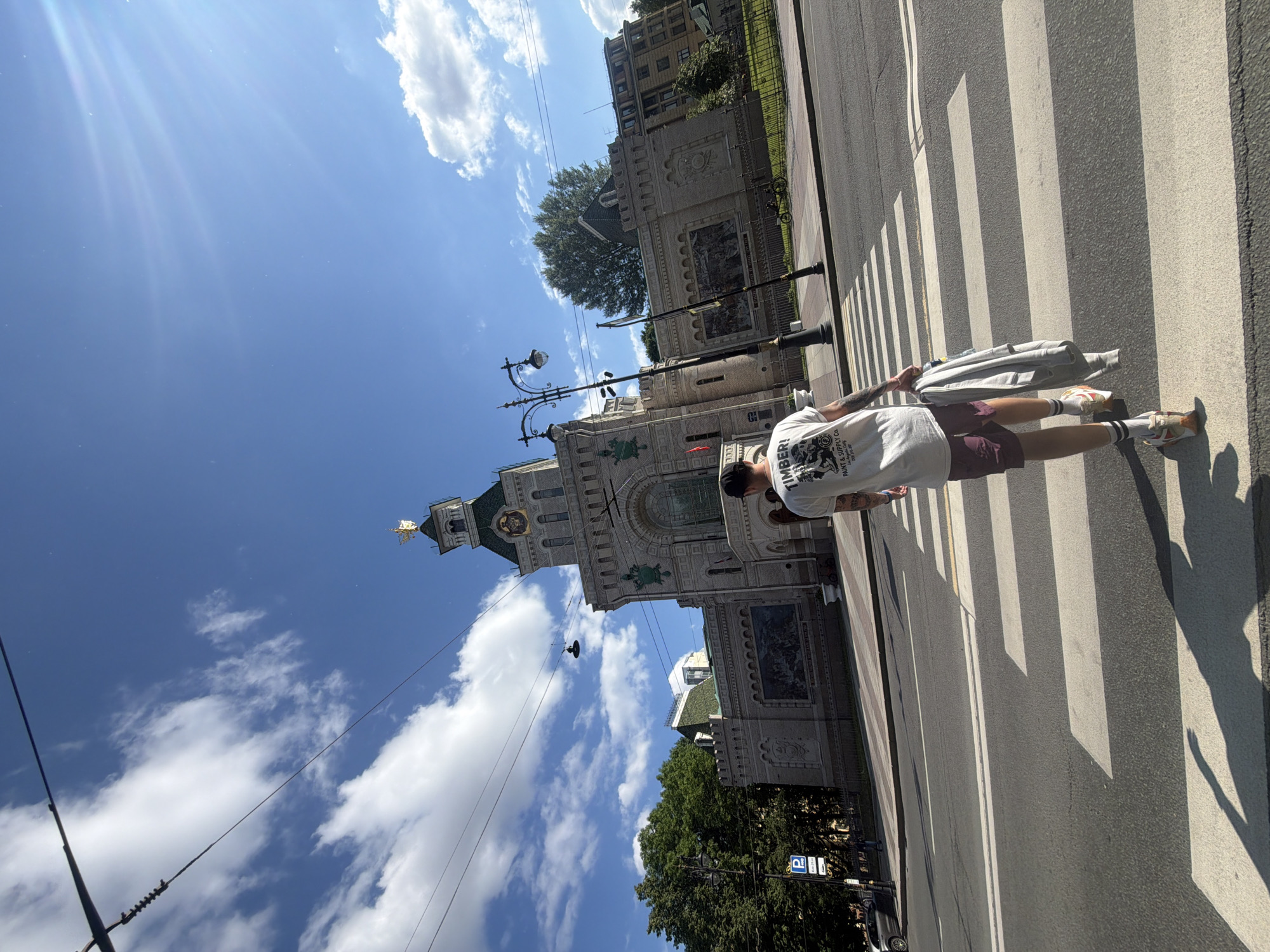 A full shot captures a young man walking away from the camera across a crosswalk. He is wearing a white t-shirt with graphics, burgundy shorts, white socks, and sneakers. He carries a grey hooded jacket over his left arm. The man is positioned slightly right of the frame's center. Behind him, a large, ornate building with a green roof and a golden spire dominates the background. The architecture is detailed, featuring murals and decorative elements. The building is situated on the other side of a street, with a grassy area and trees visible beyond it. Overhead, power lines and a street lamp are visible against a bright blue sky with scattered white clouds. The scene appears to be a sunny day in a city. This building is the Church of the Resurrection of Christ, also known as the Church of the Savior on Spilled Blood, located in Saint Petersburg, Russia.