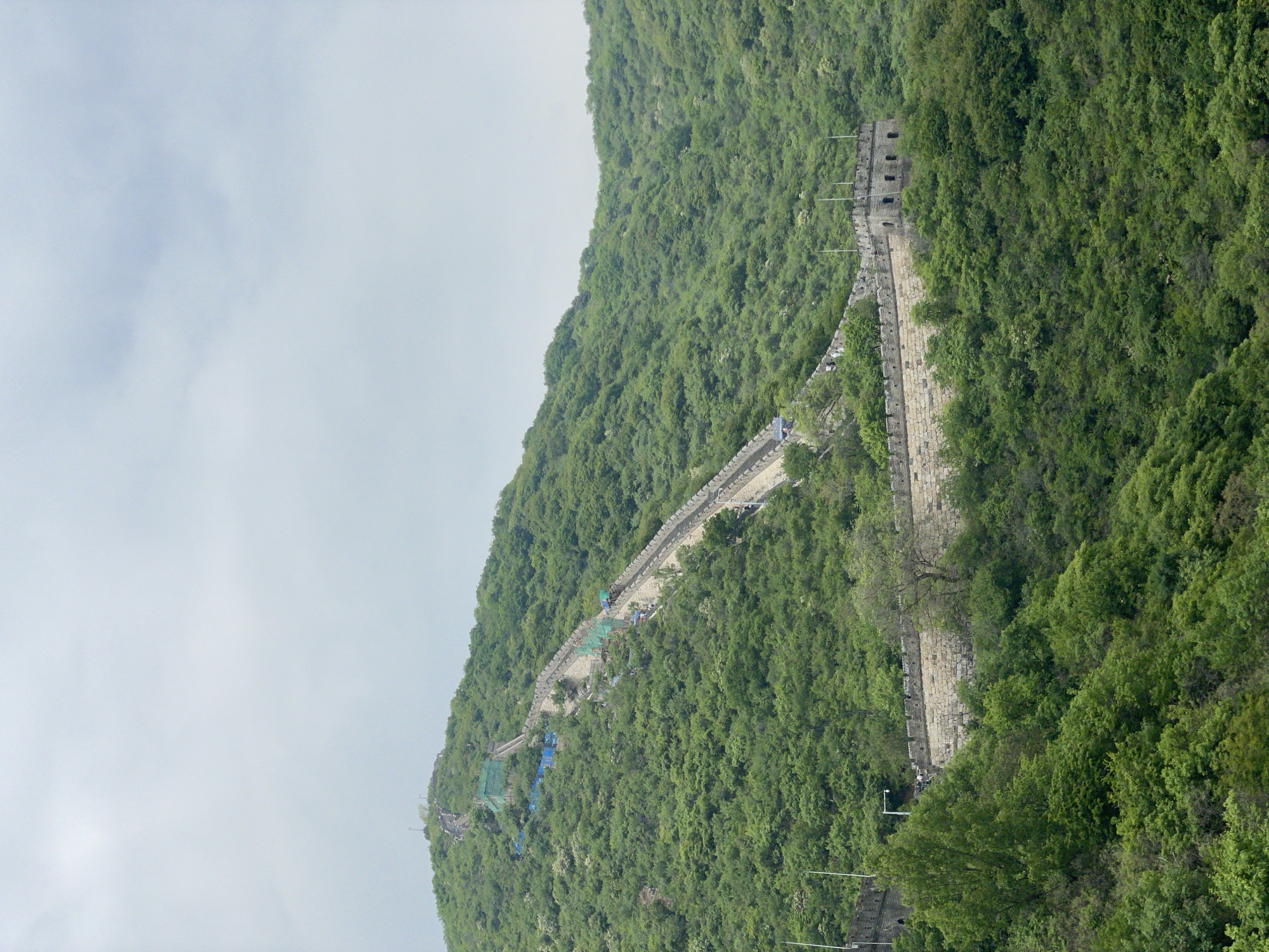 The image shows a section of the Great Wall of China, located in the Huairou District. The wall is constructed of stone and follows the ridgeline of a heavily wooded mountain. The foliage appears dense and green. The sky is overcast. The wall itself shows signs of disrepair. There are some small groups of people visible walking along the wall. Along one stretch, construction or repair work is underway as blue tarps are visible. Overall, the scene depicts a historic landmark integrated into its natural surroundings.