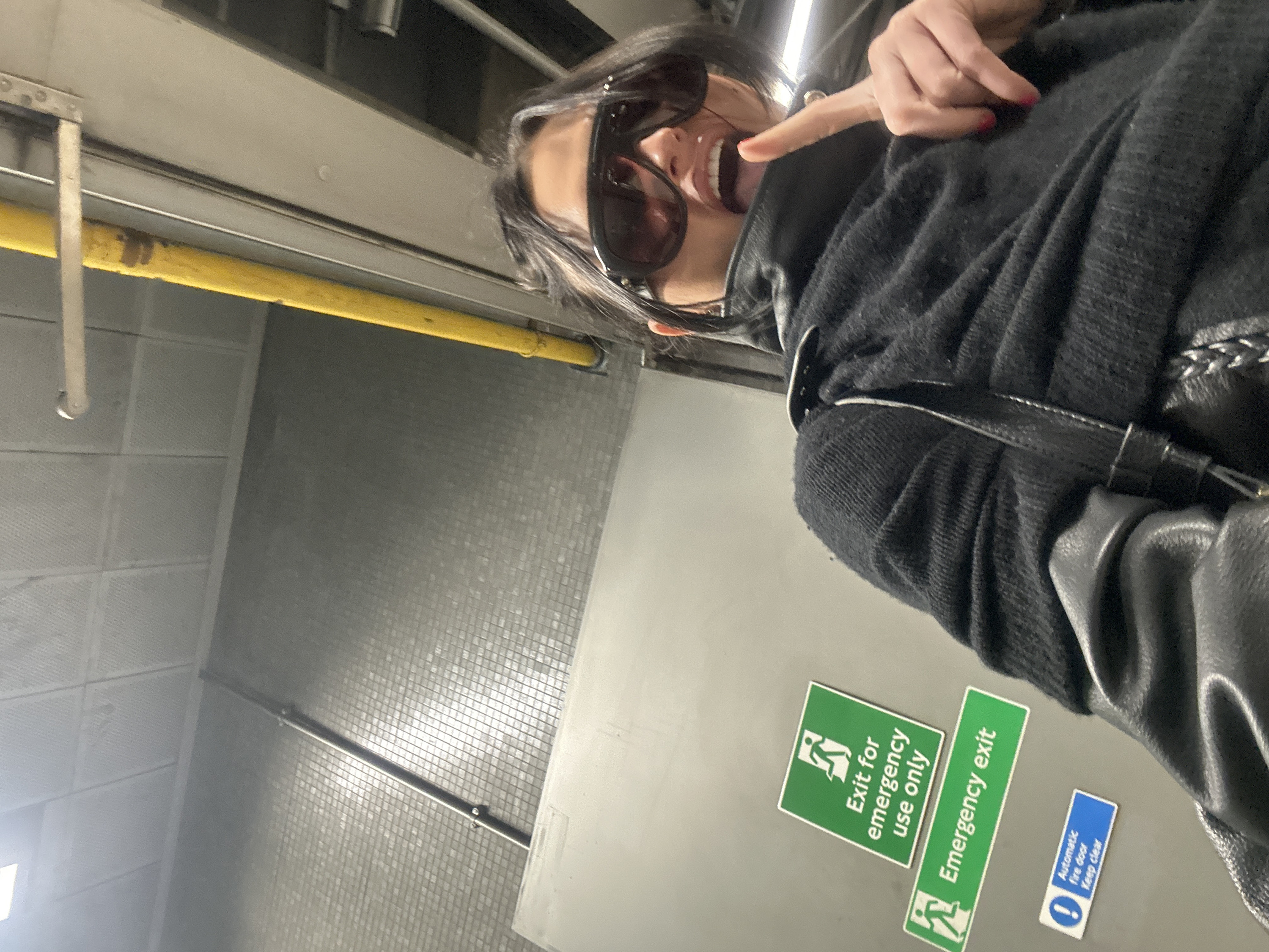 The image depicts a female individual in a public indoor setting, likely within the London Underground system or a connected passage. The subject, in the foreground, wears large dark sunglasses, a dark textured top, and a black leather jacket. Her mouth is open, tongue slightly extended, and her right index finger is pointed upwards. Red nail polish is visible on her fingernails. The background features a light grey tiled wall. Mounted on this wall are two green emergency exit signs, one stating "Exit for emergency use only" and the other "Emergency exit," both featuring a running figure pictogram. Below these, a blue sign reads "Automatic fire door Keep clear." A yellow-sleeved pipe or conduit is visible vertically along the right side of the frame, extending from the bottom to the upper right. A metallic horizontal bar or railing is also present in the upper right. The ceiling consists of square panels, illuminated by an overhead light source in the upper left. The environment's characteristics indicate a location in London, United Kingdom.