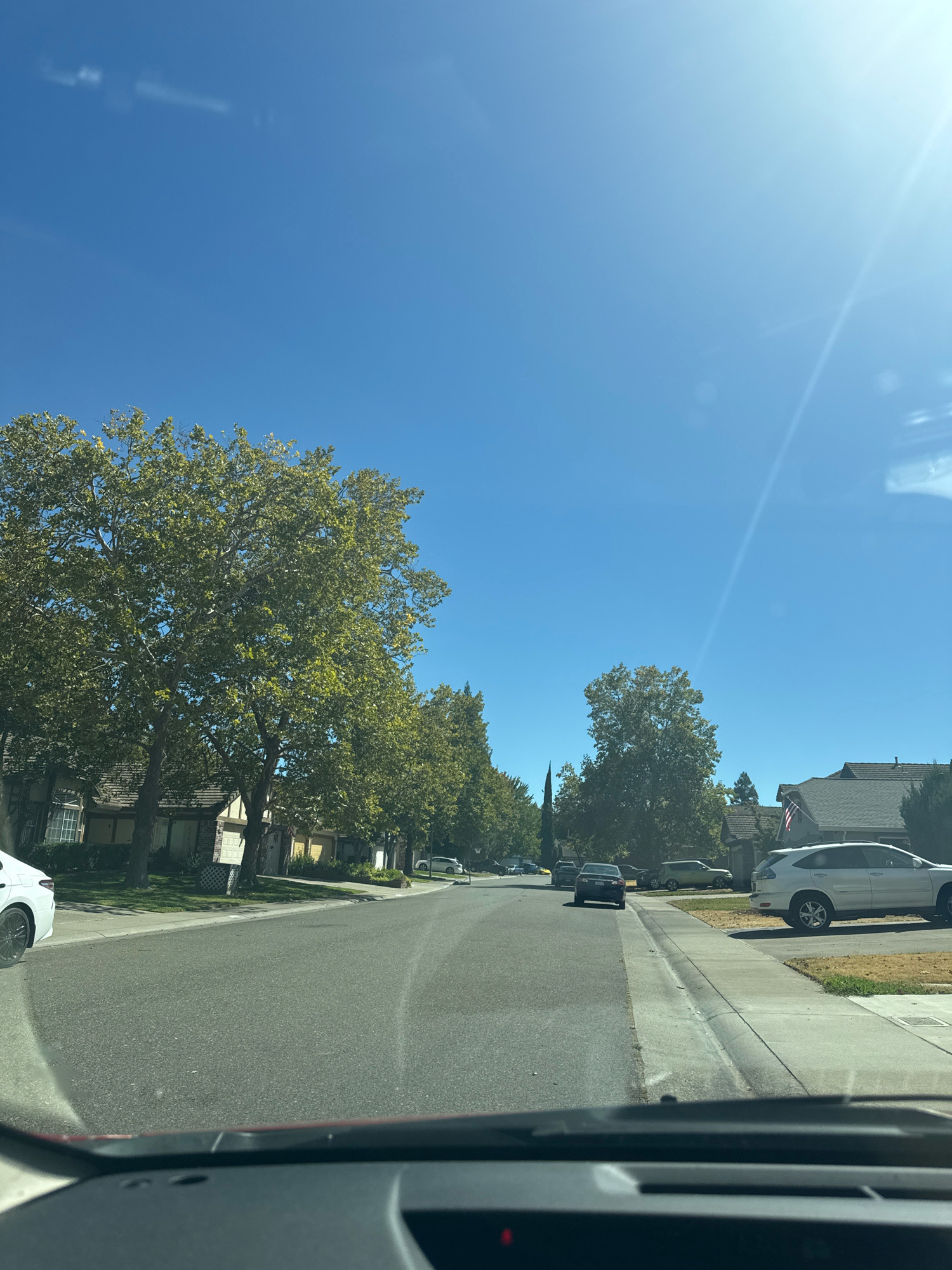 The image shows a residential street in Antelope, California, on a clear, sunny day. The perspective is from inside a vehicle, looking forward. The street is paved and lined with sidewalks and lawns. Houses with pitched roofs and visible driveways are situated on either side of the street, interspersed with mature trees that have green foliage. Several cars are parked on the street and in driveways, including a white SUV, a dark sedan, and a lighter colored sedan. The sky is a bright, clear blue with a few faint wisps of clouds and lens flare from the sun. An American flag is visible on the front of one of the houses.