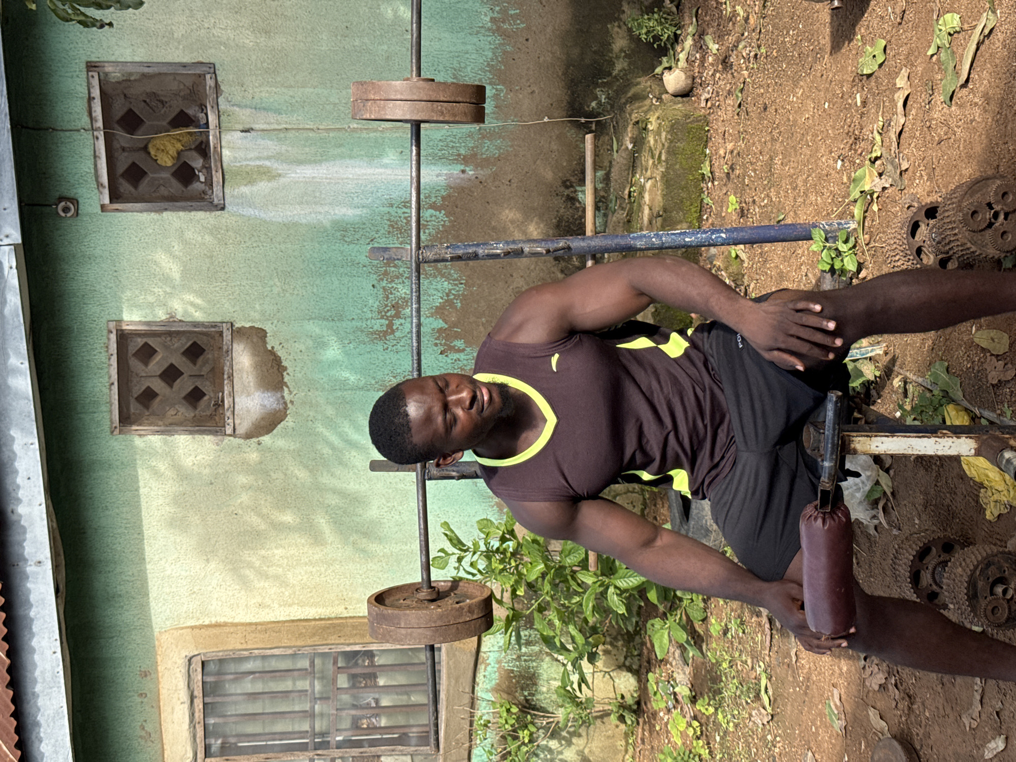 In Akwanga, Nigeria, a Black man with dark skin, a muscular build, and short dark hair is seated on a weightlifting bench in an outdoor setting. He is wearing a dark brown sleeveless athletic shirt with a neon yellow trim around the neck and armholes, and black athletic shorts. His eyes are closed, and he appears to be resting or concentrating.
The bench is old and appears to be made of metal with a dark brown padded seat. Beside the bench, on the ground, are various repurposed metal objects that seem to be part of the weightlifting equipment, including what looks like gears and parts of an engine. A barbell with heavy circular weights is positioned behind the man, resting on supports.
The background consists of a weathered, light green building with a corrugated metal roof. The building has two small square windows with decorative blockwork. The area around the bench is a dirt and gravel surface with some sparse green vegetation and fallen leaves. The overall impression is a functional, albeit rustic, outdoor gym setup.