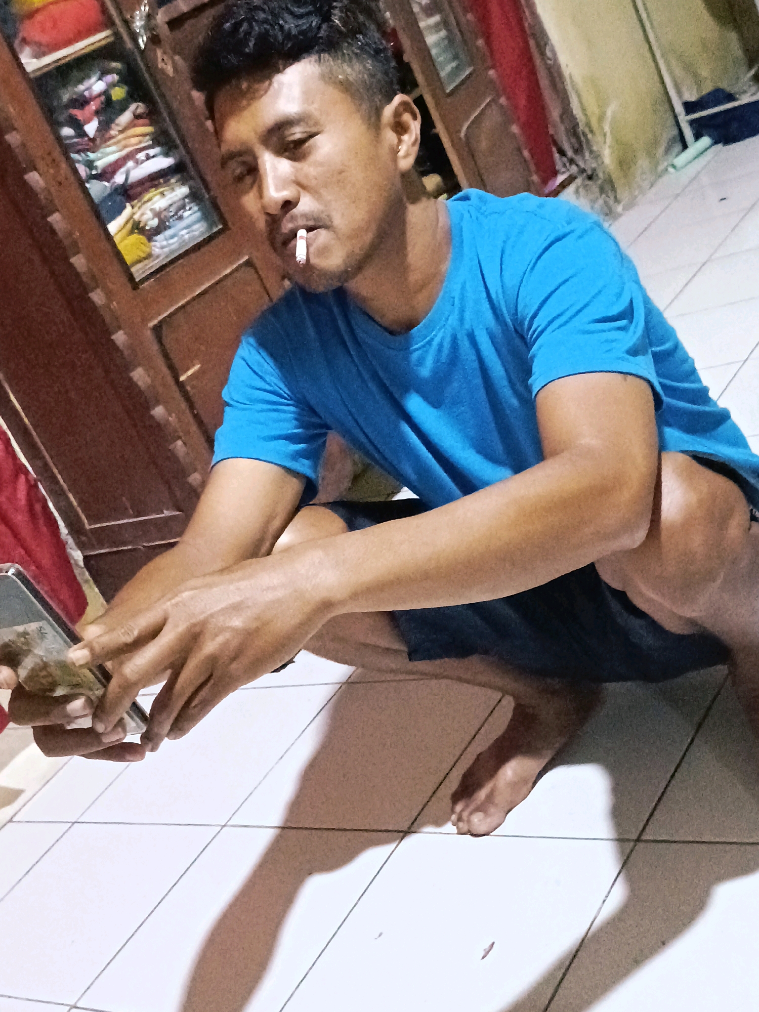 The image depicts a man crouching on a tiled floor indoors, holding a smartphone in his left hand. A lit cigarette is visible in his mouth. He is wearing a blue t-shirt and dark shorts. In the background, a wooden cabinet filled with clothes is partially visible, as is a wall painted in muted tones. The lighting suggests an indoor environment, possibly a home. The scene appears to be located in Jatitengah, Indonesia, based on the provided context.
