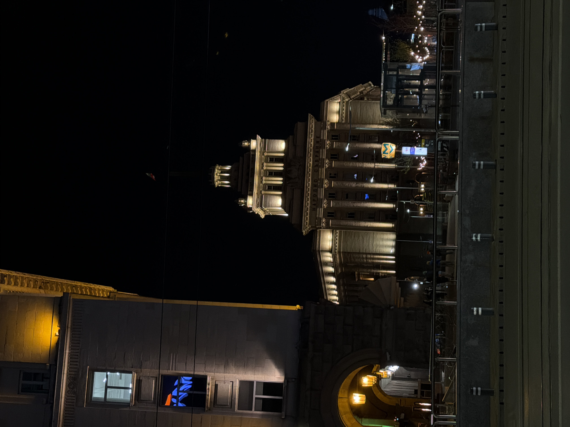At night, the image captures a view of architecture in Sofia, Bulgaria. The primary structure is a grand, ornate building with lit columns, likely a governmental or cultural institution, suggesting the location is in a central area of the city. Below the building, there is another building with lit windows; a neon blue shape is visible in one of the windows. In the foreground to the right, a metal structure is seen, with white stripes on the posts. A street scene is also captured between buildings, lined with decorative lights.