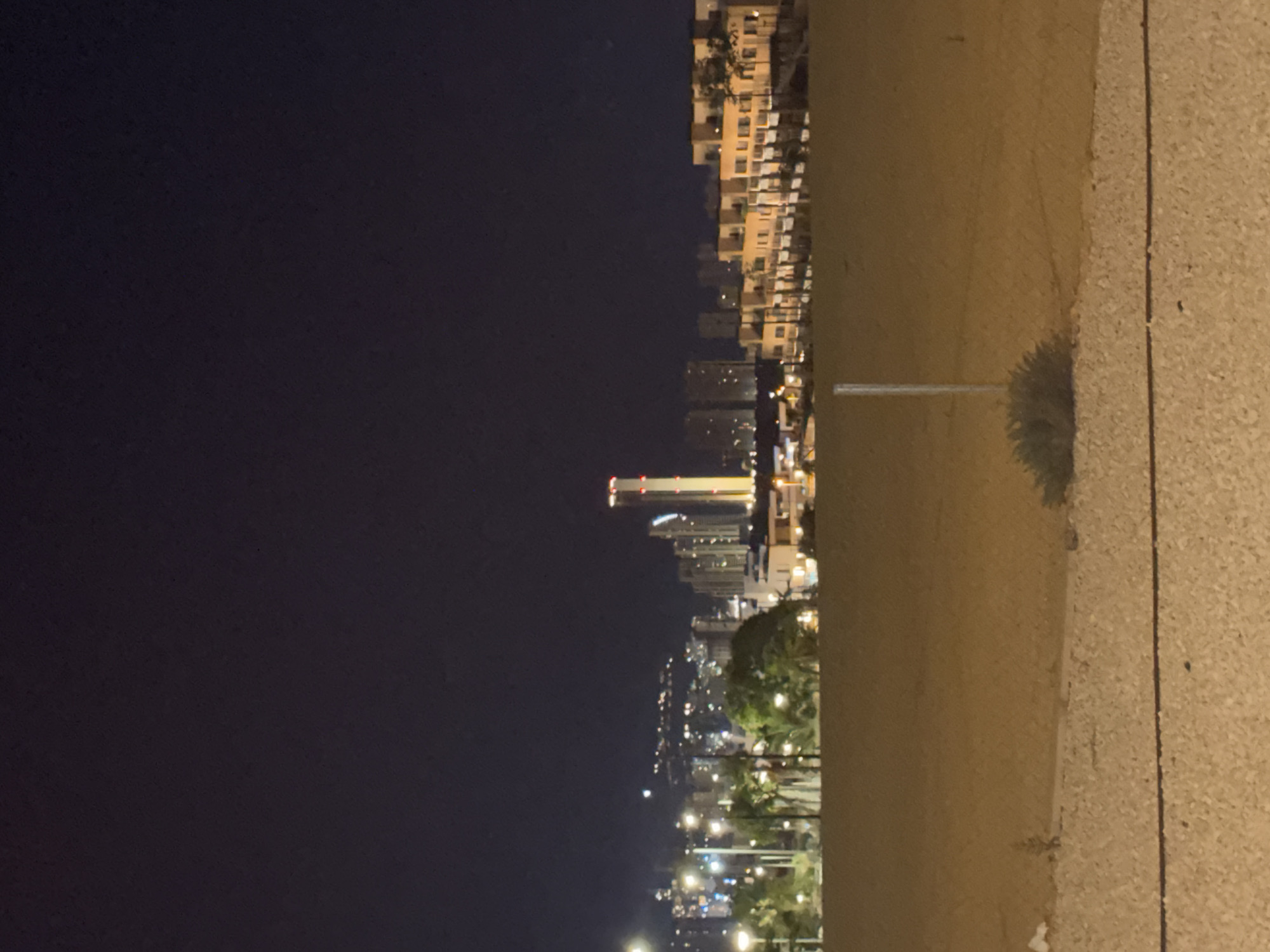 A night-time, wide-angle, eye-level shot captures a cityscape in Finestrat, Spain. The foreground features a sandy, uneven terrain with scattered small plants and a grey gravel-like surface, delineated by a thin dark line. In the mid-ground, a band of low-rise buildings with illuminated windows and balconies stretches across the frame, constructed of a light-colored material. Beyond these, a more distant cityscape reveals a cluster of taller buildings and a prominent, illuminated skyscraper. The sky above is dark, indicating night.