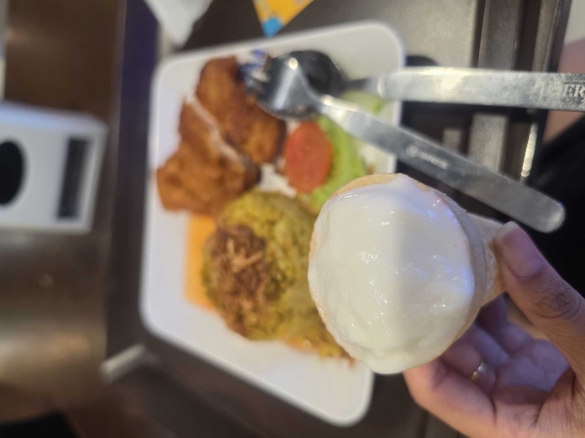 A high-angle, slightly blurry photograph shows a meal on a white plate, with a hand holding a cone filled with white substance in the foreground. In the center of the frame, the white plate contains fried chicken pieces, yellow rice, lettuce, and a sliced tomato. Silver utensils, a fork, and a knife, are placed on top of the food. The hand, presumably holding the cone, belongs to a person with dark skin. The background appears to be a dark surface. Based on the provided context of Bangkok, Thailand, this is most likely a meal served onboard a Thai Airways flight and the ice cream is the desert.