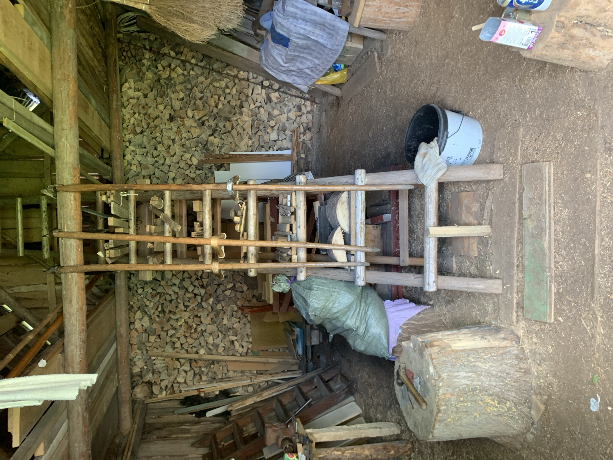 The image depicts the interior of a rustic wooden structure, possibly a workshop or woodshed, located in Відзаўскі сельскі Савет, Belarus. The floor is covered with dirt and wood shavings. A large wall of neatly stacked split firewood occupies the entire rear of the building. In the center, a multi-tiered wooden rack or stand, constructed from logs and planks, holds numerous small pieces of cut wood and several long-handled tools, possibly chisels or gouges, secured with cordage. Raw split log segments are also visible on a lower shelf of this rack.
To the left, a large tree stump serves as a chopping block or workbench, with a hammer and a wedge-like tool resting on its surface. Behind the stump, a voluminous green woven sack sits atop a small wooden stool or crate draped with a light pink cloth. Further left, a wooden ladder leans against a plank wall, surrounded by various planks and timbers.
To the right, a significant quantity of dried plant material, possibly brooms or hay, hangs from an overhead wooden beam. Beneath this, a makeshift resting area is fashioned on what appears to be a large box or bench, covered with a grey and blue striped blanket. A person is partially visible underneath the blanket, with their head and a blue garment discernible. Adjacent to the resting area, a yellow plastic bag and a plastic bottle are on the floor. In the immediate foreground to the right, a white and black bucket with a white cloth draped over its rim is visible. Further to the right, an open pink and white cardboard package (with "ЧРЕЗМЕРНОЕ" text) and a metal can rest on a small wooden stump.
Overhead, exposed wooden beams and planks form the ceiling structure. A white cloth or garment hangs from a central beam on the left side of the frame. Several green-painted wooden planks lie on the floor in the foreground, possibly serving as steps or floor protection.