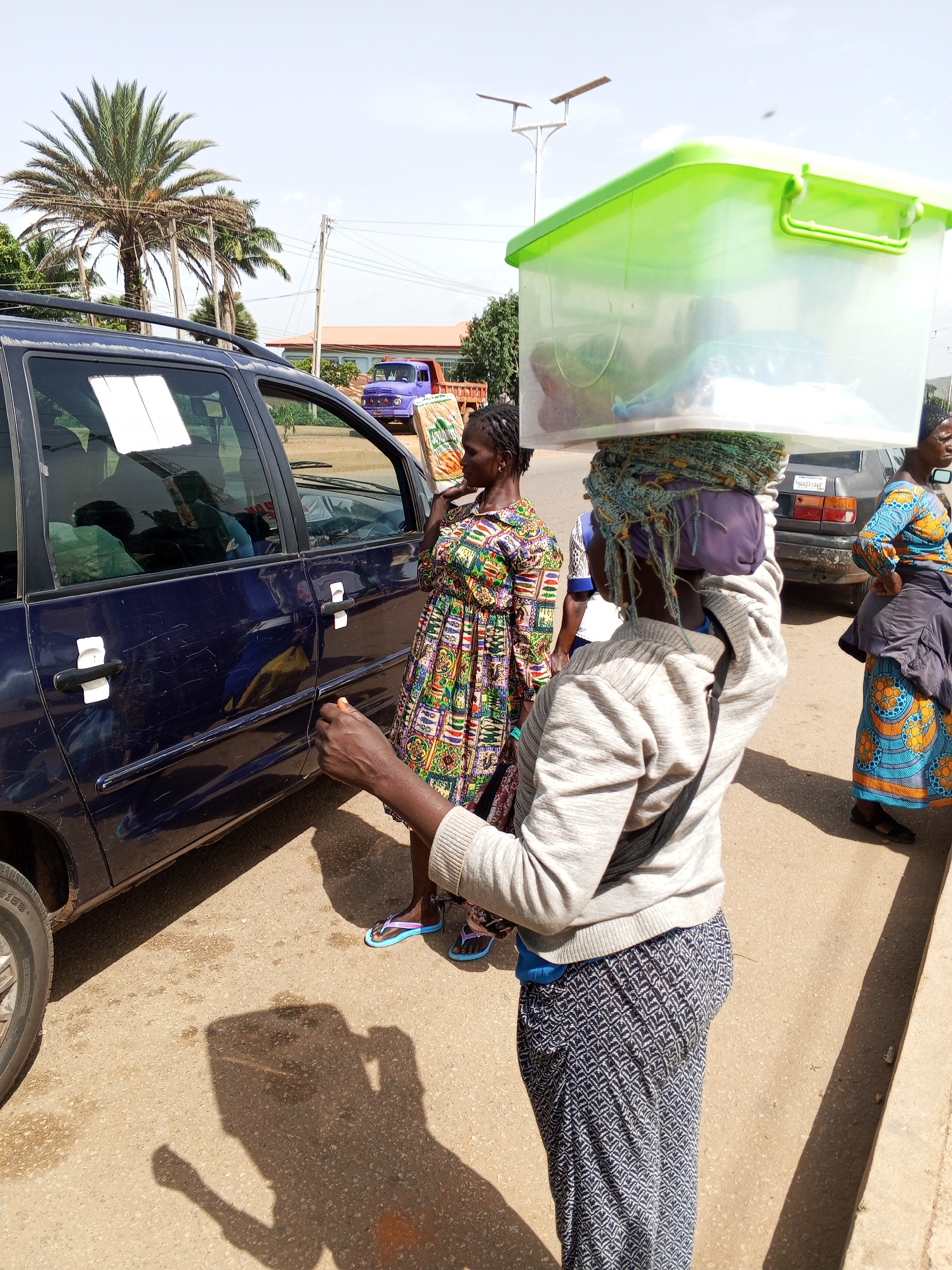 A woman in a gray sweater and patterned pants balances a large, translucent green plastic container filled with items on her head, supported by a bundle of fabric. She stands on a paved road next to a dark blue minivan. To her left, another woman in a colorful dress holds a bag of bread. In the background, a purple truck and other vehicles are visible. The scene appears to be in an urban area, possibly Bukuru, Nigeria, given the presence of palm trees and buildings consistent with the region. The lighting suggests it is daytime.
