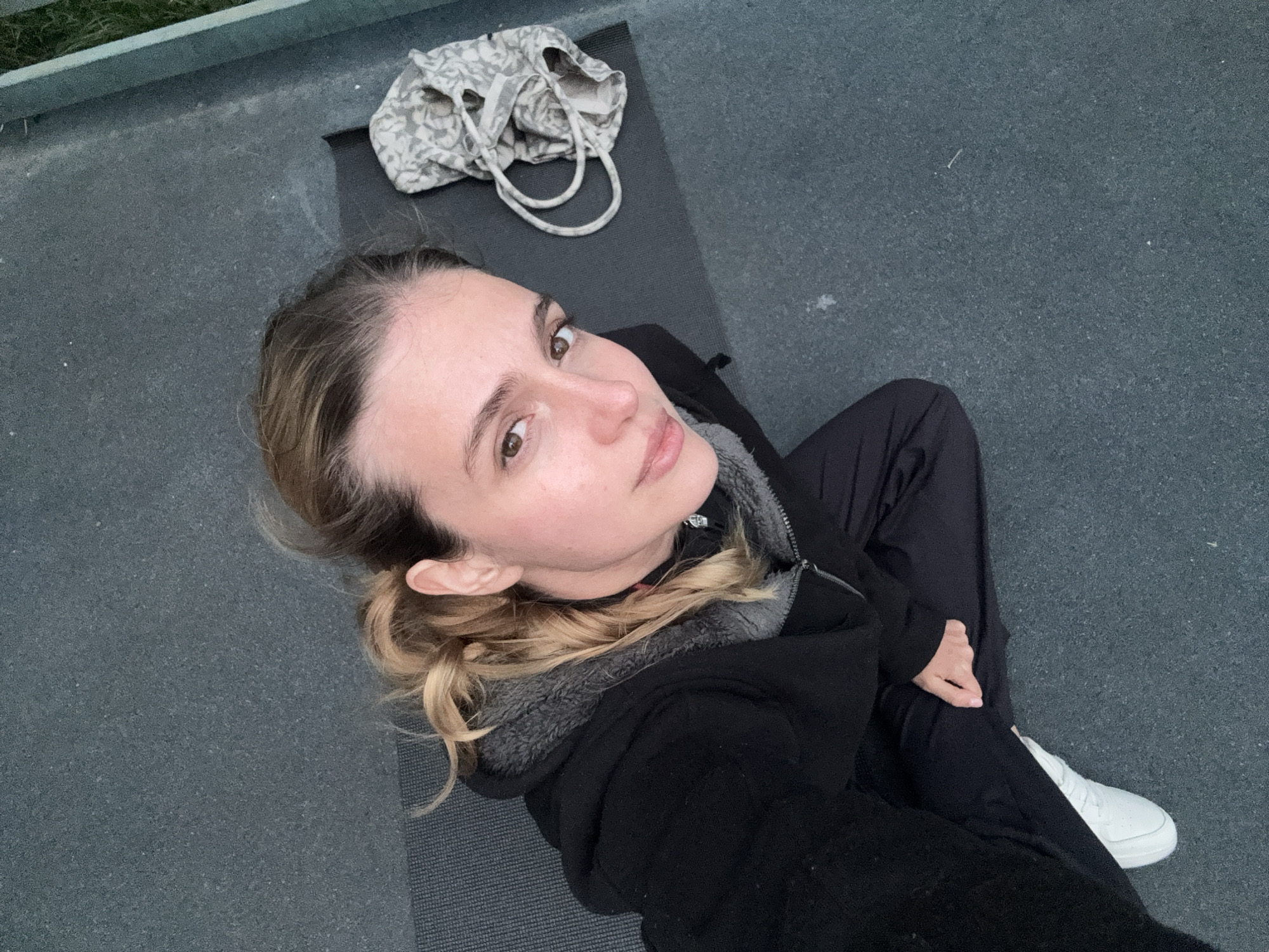A young woman is depicted in a high-angle selfie or portrait, seated outdoors on a dark grey, textured surface, possibly concrete. She has light brown hair tied back and brown eyes, looking directly at the camera with a calm, neutral expression. She is wearing a black jacket with a grey fleece lining and black pants, along with a white sneaker on her right foot.
She is positioned on a dark grey yoga mat or similar padded surface. Behind her on the mat, there is a light-colored, possibly off-white or light grey, patterned bag with handles. In the upper left background, a light-colored fence or railing is visible, with a hint of green foliage beyond it. The soft, even lighting suggests the photograph was taken during an overcast day, or possibly during early morning or late afternoon. The setting implies an urban outdoor space, such as a rooftop or terrace, located in Tbilisi, Georgia. There is no visible text in the image.