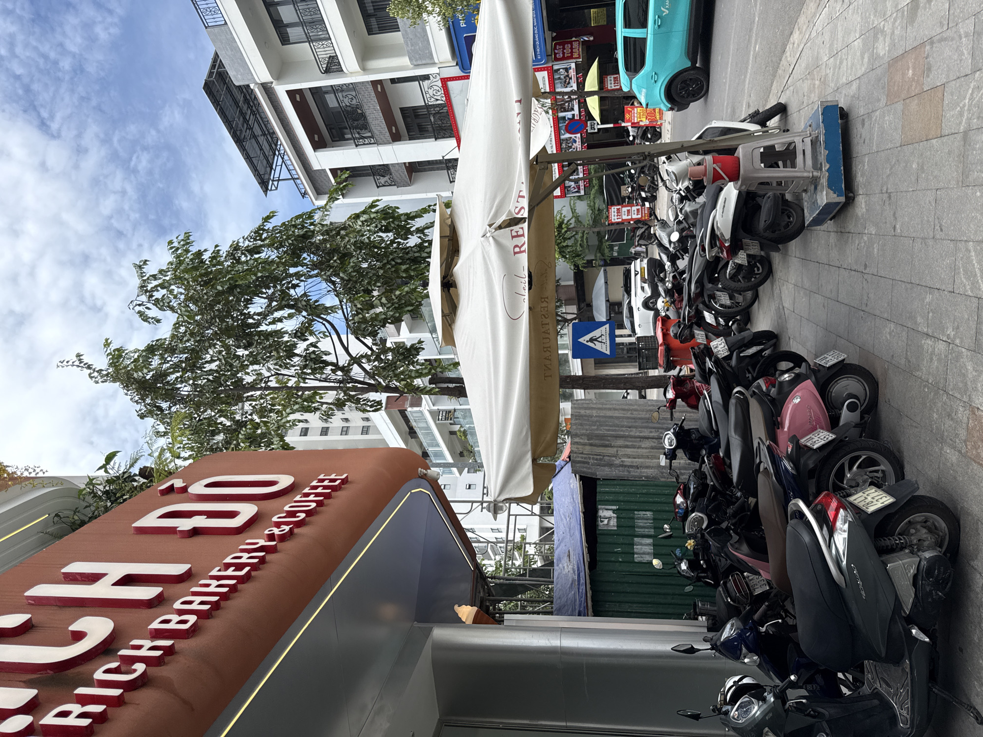 The image shows a street scene in Nha Trang, Vietnam, with a focus on a bakery and coffee shop named "RICH BAKERY & COFFEE" with Vietnamese characters. The bakery sign is prominently displayed in the upper left corner of the image, featuring large, red, three-dimensional letters. In front of the establishment, a row of parked motorcycles and scooters lines the sidewalk. A large white outdoor umbrella, seemingly from a restaurant, is set up in front of the bakery, casting a shadow over some of the parked vehicles. Beyond the umbrella, more parked motorcycles and a turquoise car are visible on the street. Buildings, including a taller, modern white building with balconies, rise behind the street scene. Trees and a partially constructed structure with green corrugated metal walls are also present. The sky is partly cloudy.