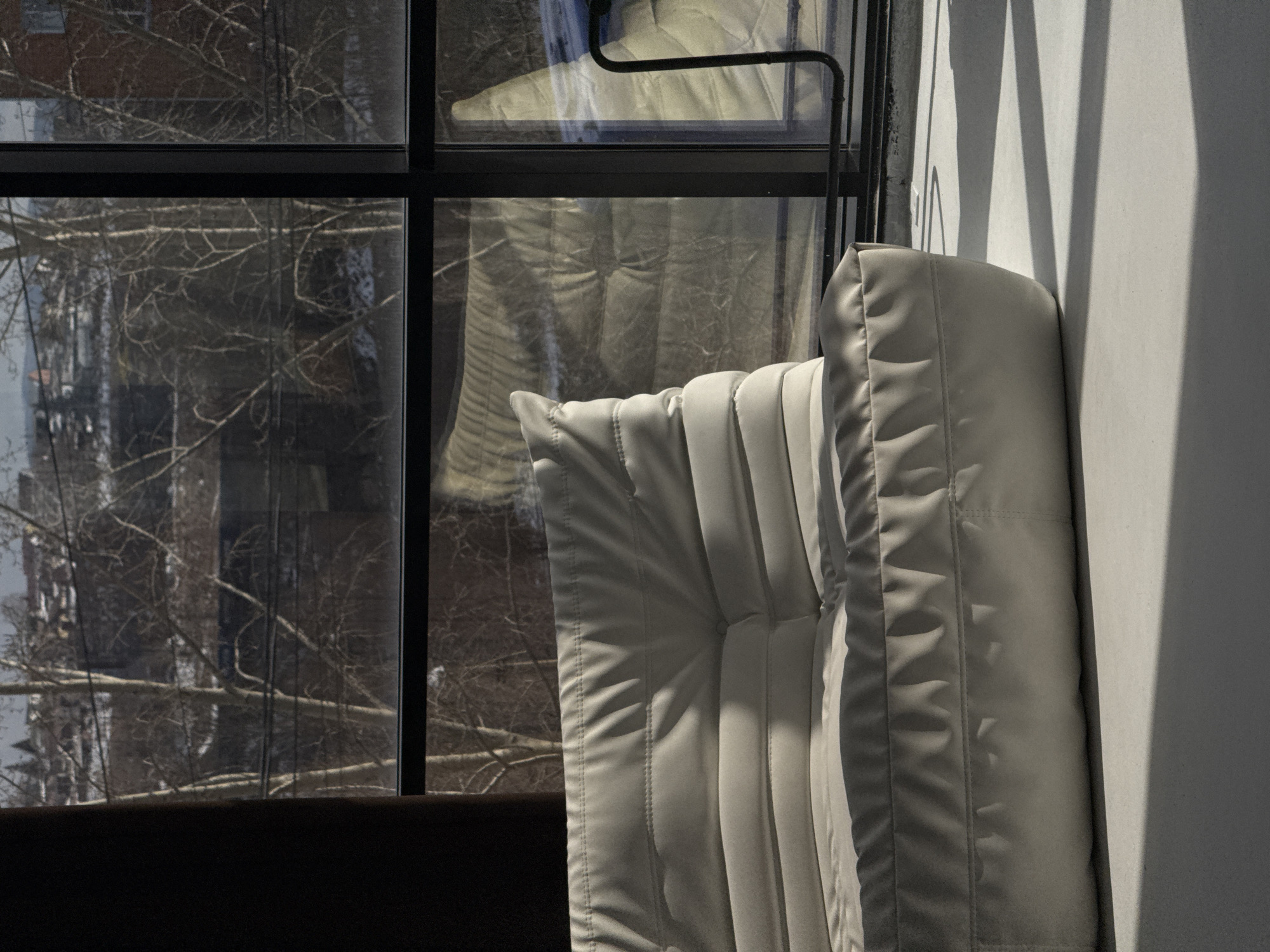 A photograph captures a partially visible, white quilted cushion resting against a white wall inside a room with a large window. The window, framed in black, offers a view of bare trees and indistinct buildings, possibly in Krasnoyarsk, Russia. A portion of dark-colored furniture is visible at the bottom of the frame. The light casts shadows along the wall, enhancing the quilted pattern of the cushion. A slender black metal object is attached to the top of the window frame.