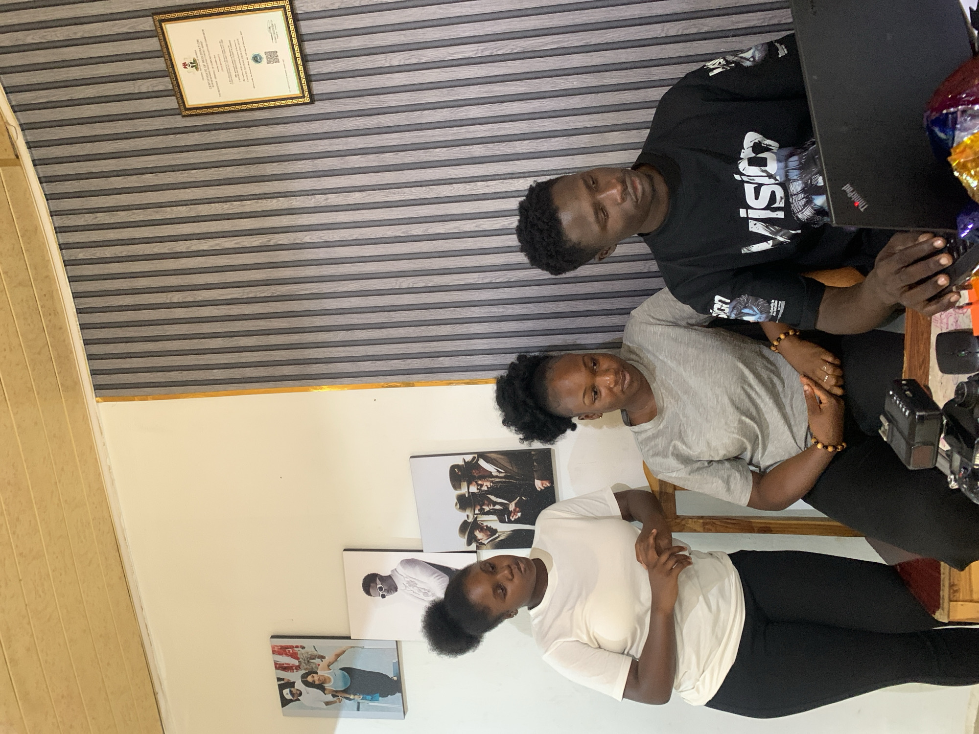 Three young adults are situated indoors. A man is seated at a table, utilizing a laptop. Next to him sits a woman in a gray t-shirt and black leggings. Below, another woman, wearing a white t-shirt and black leggings, stands. There are framed photographs on the wall. A document in a frame is on the wall. The picture appears to be taken in Jos, Nigeria.