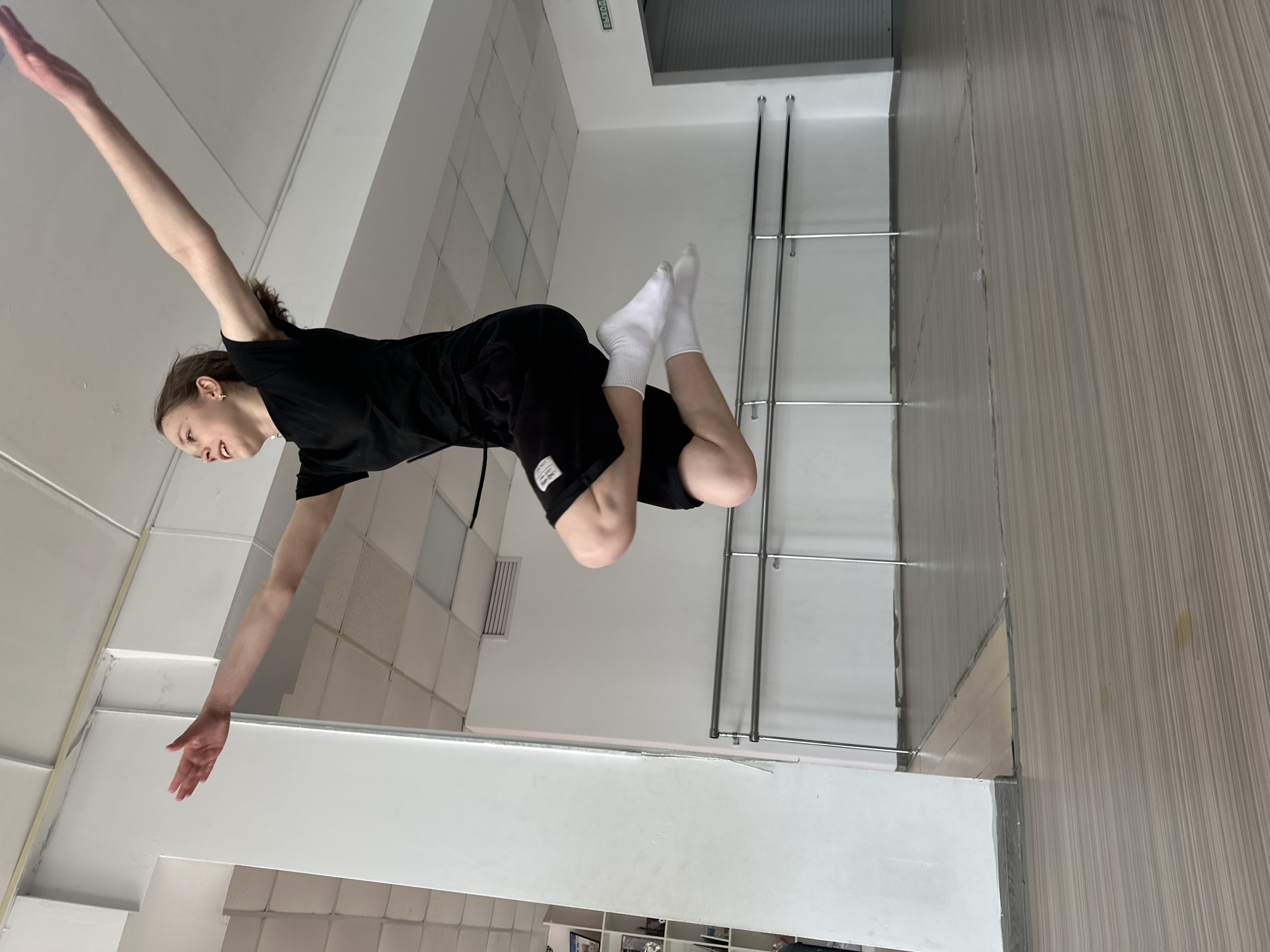 A person is suspended horizontally in a room with tiled walls and a wood-paneled wall. The person has one arm extended upwards and the other extended downwards. The person is wearing black shorts, a black shirt, and white socks. Two horizontal metal bars are visible on the wall behind the person's legs. The location is likely Votkinsk, Russia.