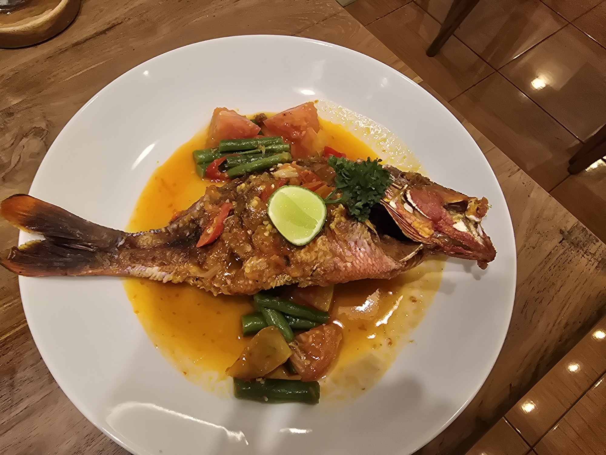 On a wooden table with a tile floor nearby is a white plate of food. The food is a whole fried fish with a red head and a brown tail, covered in an orange sauce with green beans and tomato pieces. A slice of lime and a sprig of parsley sit on top of the fish. A small wooden bowl is in the upper left corner of the frame. The scene is in the city of Unknown, Unknown.