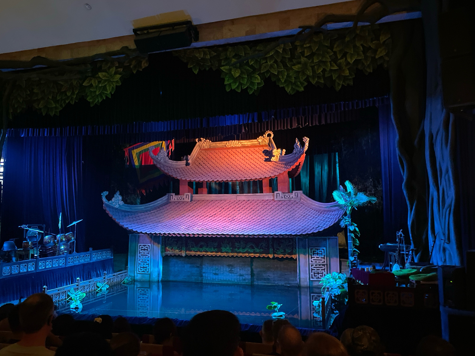 The image captures a water puppet show performance at the Thăng Long Water Puppet Theatre in Hanoi, Vietnam. The stage features a traditional Vietnamese architectural structure with a curved, tiled roof, illuminated with pink and purple lights. Below the roof, a backdrop depicting dragons and intricate patterns is visible. The performance area is a shallow pool of water, reflecting the stage lights and the surrounding scenery, which includes artificial trees and blue curtains.
On the left side of the stage, a drum set and other musical instruments are placed on a raised platform adorned with decorative railings. To the right, more musical instruments, including what appears to be a keyboard and string instruments, are set up on a platform. Several small green plants are scattered around the stage, some floating on the water.
In the foreground, the backs of an audience are visible, seated in rows of chairs, suggesting they are watching the performance. The overall lighting is dim, with spotlights focused on the stage, creating a theatrical ambiance. The scene is captured from a perspective within the audience, looking towards the stage.
