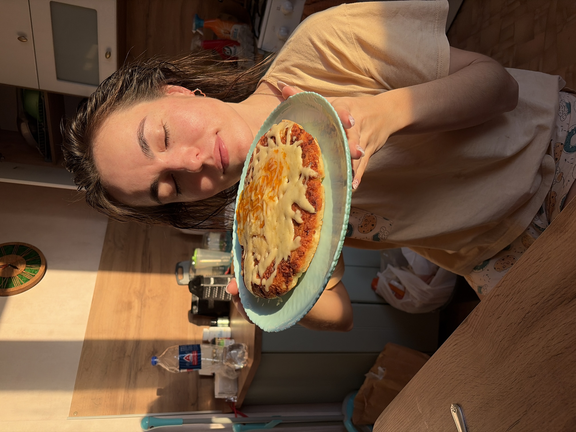 A domestic kitchen setting in Izhevsk, Russia, is illuminated by direct, low-angle sunlight casting distinct shadows. A female individual is positioned centrally, holding a light blue plate with a scalloped edge. Her eyes are closed, and her head is slightly tilted, suggesting she is inhaling the aroma of the food on the plate. Her dark hair appears damp, and she wears a light beige t-shirt with patterned pajama shorts. Small rose-gold hoop earrings are visible.
The plate holds a round, baked food item with a reddish-orange base heavily covered with melted, golden-brown cheese. In the background, a light-colored wall features a wooden-patterned backsplash. A white upper kitchen cabinet with a glass insert, a gas range, and various kitchen items are visible on a countertop, including a clear plastic water bottle with a blue cap and red label, a black grater, and other containers. A light blue cleaning tool is propped against a wall. A wooden table surface is visible in the foreground, with a silver utensil resting on it. On the floor in the lower left, a brown paper bag and a white plastic bag are present.