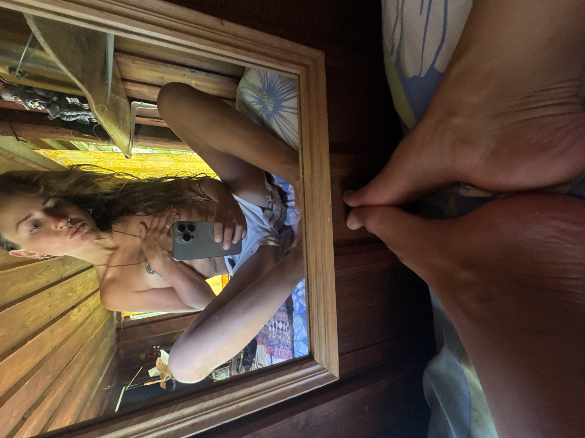 The image is a selfie taken by a young woman in what appears to be a rustic, wooden room. The woman is in the foreground, reflected in a large mirror. She is holding a smartphone to take the picture, and her gaze is directed at the phone.
The woman is bare-chested and wearing patterned underwear. Her hair is long and dark, falling over her shoulders. She has a necklace on and appears to be wearing earrings. In the reflection, her legs are crossed, and she is looking towards the camera with a neutral expression.
The room has wooden walls and ceiling, giving it a cabin-like feel. In the reflection, there are various objects visible, including a statue or figurine on a shelf and a patterned fabric, possibly a bedspread or blanket. The lighting suggests it might be daytime, but there are no specific indicators of time or weather. The overall mood is casual and personal, as if capturing a private moment. The feet of the woman are also visible in the foreground, on top of a patterned fabric.
