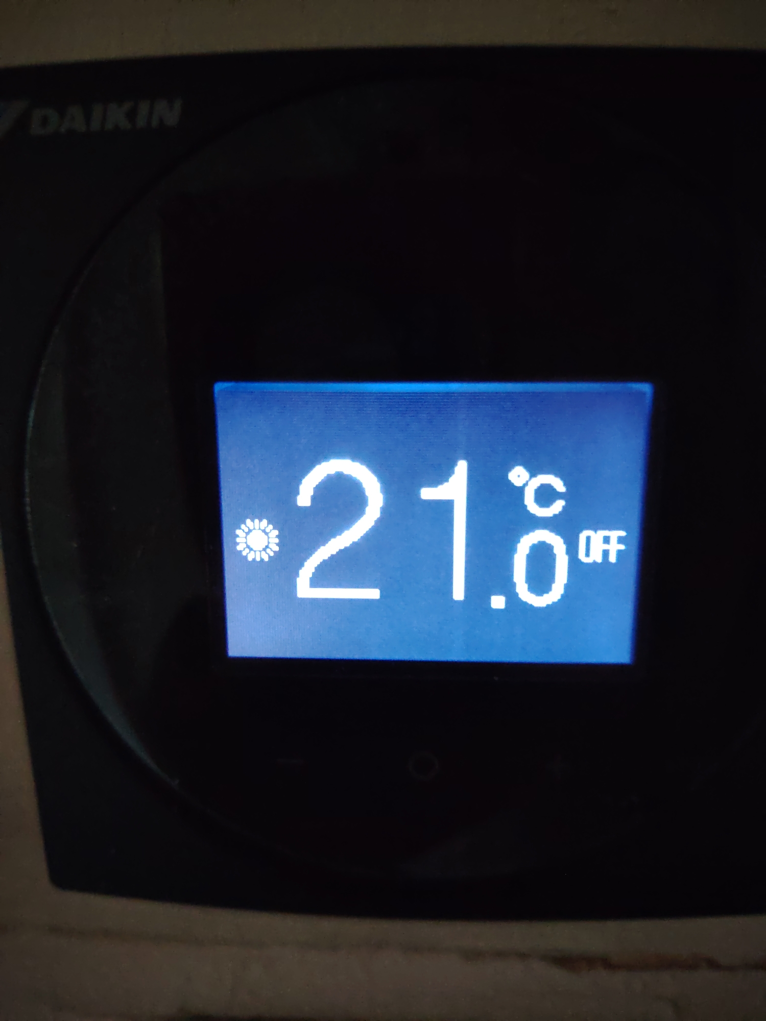 A close-up, dimly lit photograph shows a digital display on a black appliance, likely a Daikin air conditioner. The display is illuminated with blue light and shows the temperature as 21.0 degrees Celsius. A small symbol resembling a sun or flower is to the left of the temperature reading. The word "OFF" is visible to the right of the temperature. The appliance has the Daikin logo in the upper left corner. The surrounding environment is dark and out of focus, suggesting the photograph was taken indoors at night or in a poorly lit area. There is no information available to pinpoint the location within Huntingdonshire, United Kingdom.