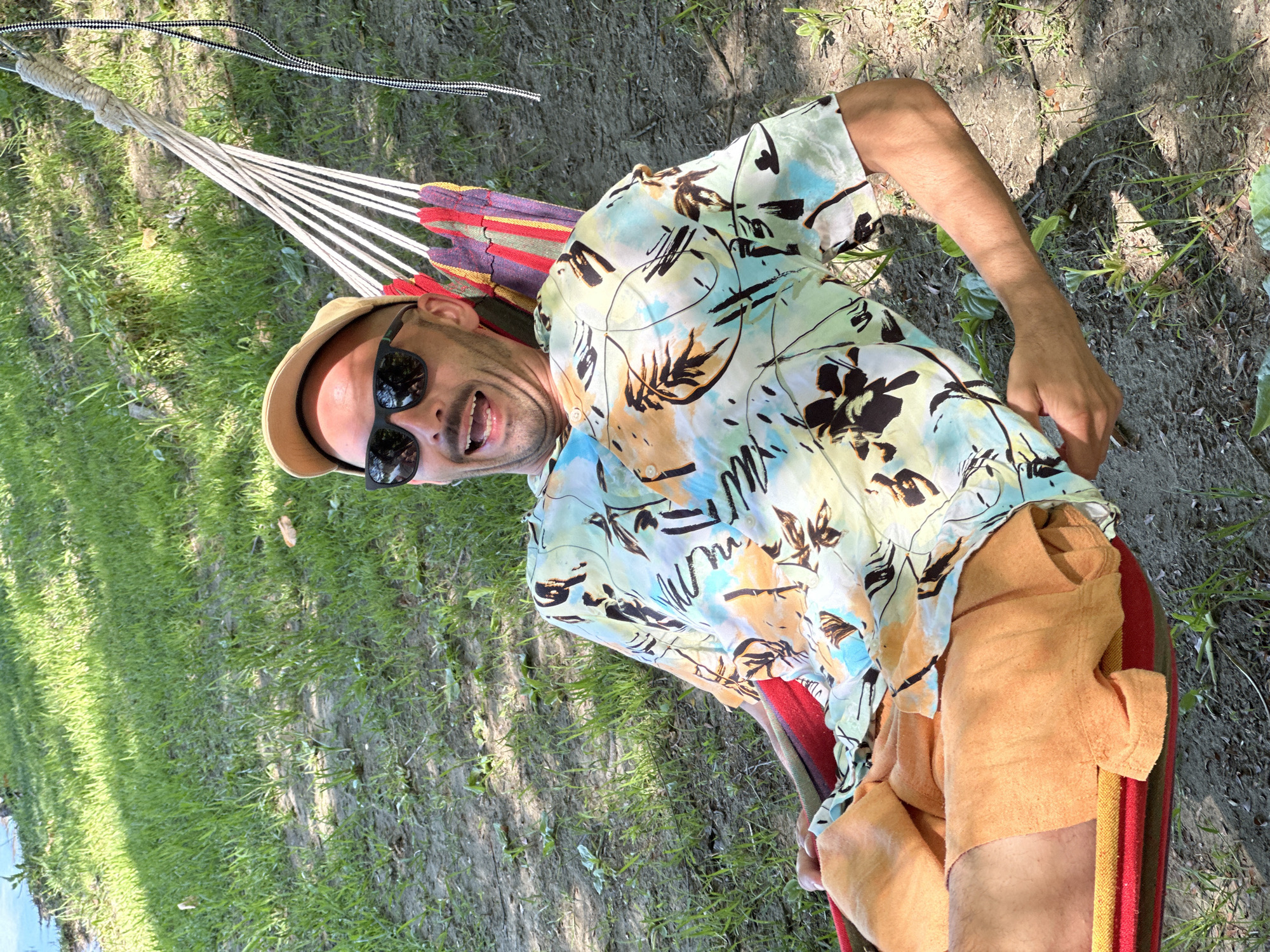 A man reclines in a hammock outdoors. He wears a patterned short-sleeved shirt, tan shorts, a tan hat, and sunglasses. He is smiling with his mouth open. The hammock is suspended between two points, and the surrounding environment includes grass and dirt. The location is Vranjina, Montenegro.