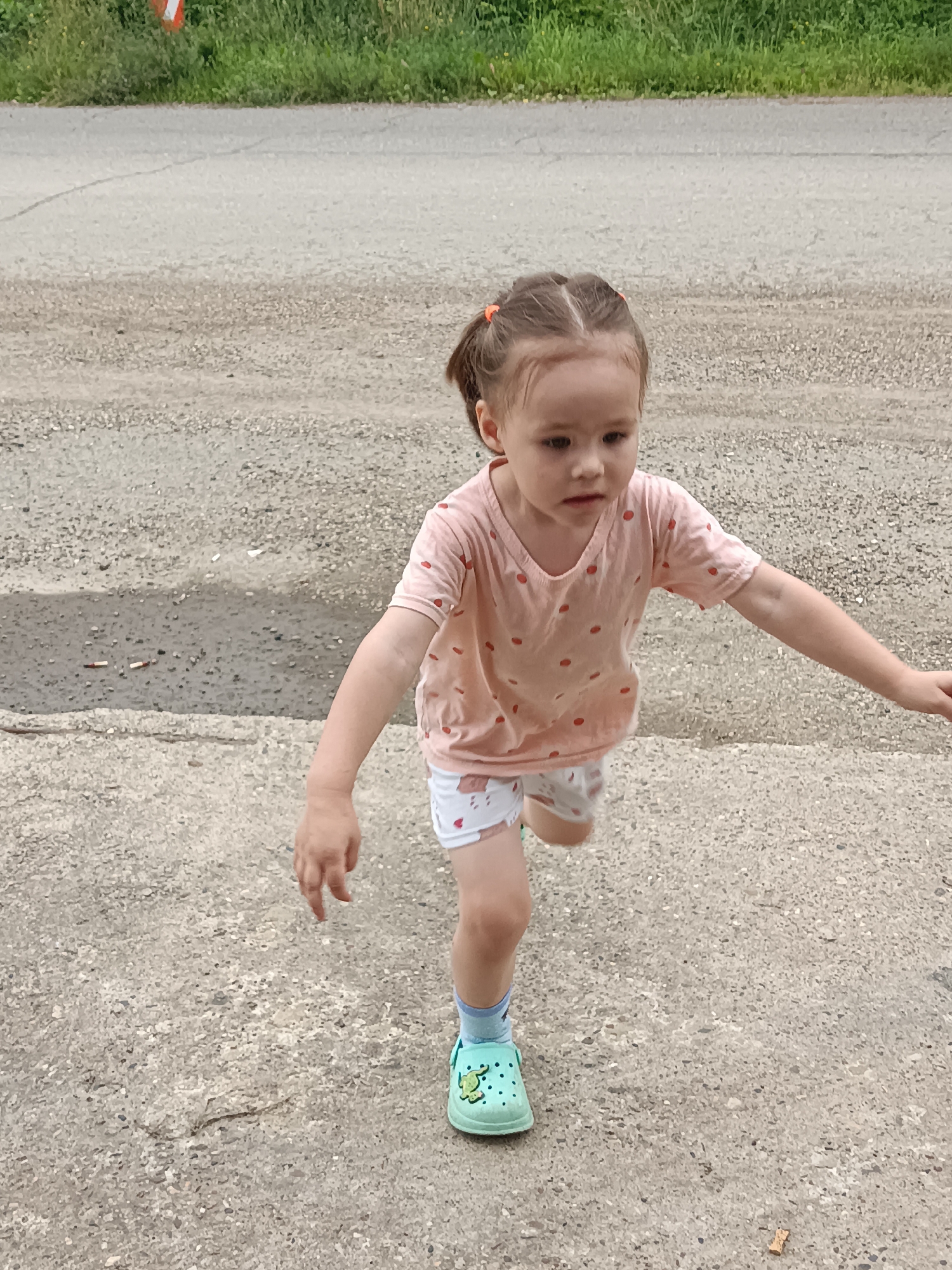 A young girl with brown hair in two pigtails tied with orange elastics is standing outdoors on a gravelly, asphalt surface. She is wearing a light pink t-shirt with small red dots and white shorts with a red and white pattern. She has on light blue socks and green clogs with a green frog design. Her right arm is extended to her right, and her left arm is bent. She appears to be in motion, possibly running or walking. The background shows a line of green grass and a grey road. The image is taken from a slightly elevated perspective looking down. There is no visible indication of the specific location within Podgorensky, Russia.
