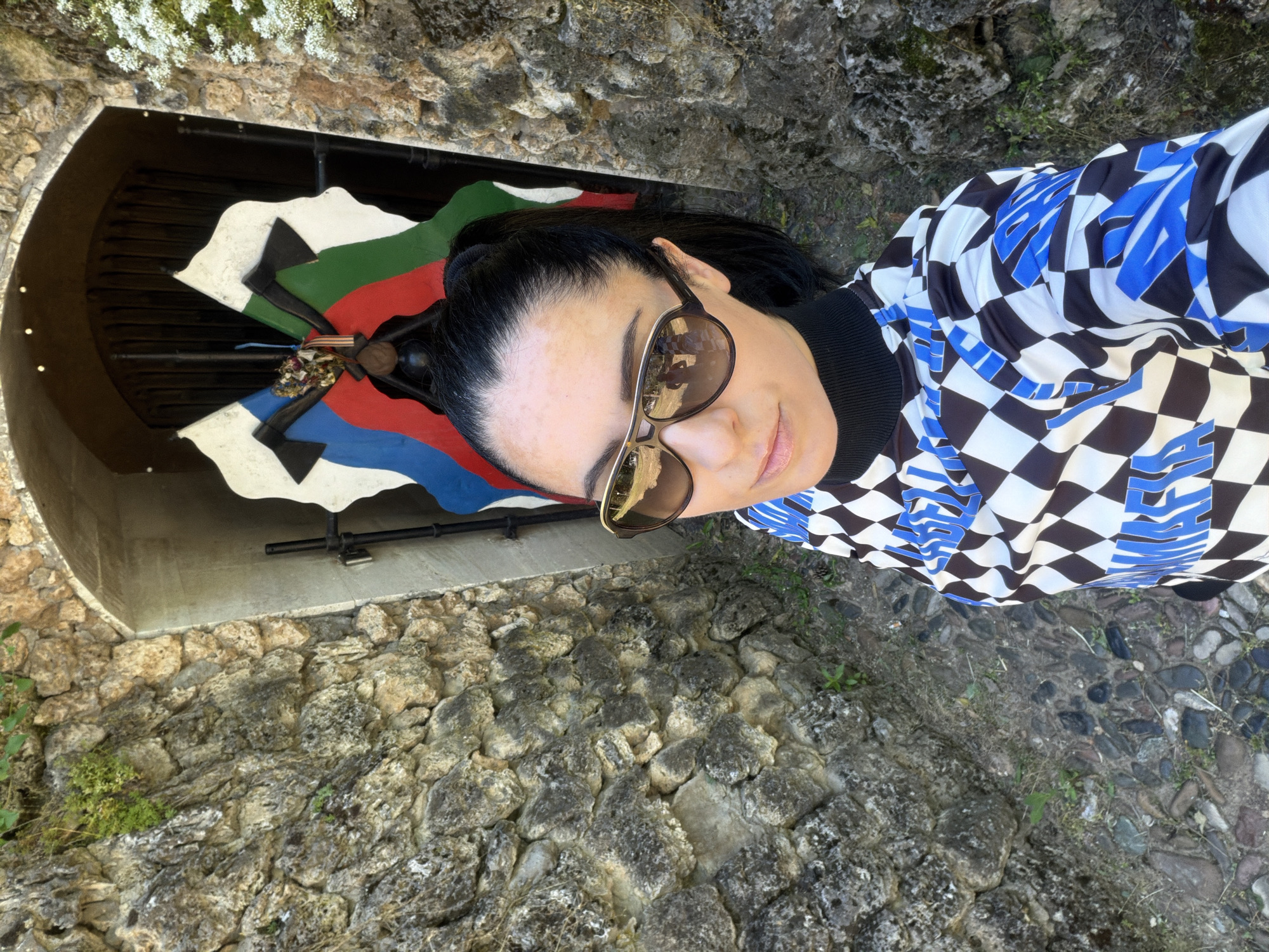A woman with dark hair and sunglasses is posing for a selfie. Behind her is the "Boat of Bones," a memorial in Pleven, Bulgaria. The memorial is built into a stone structure that resembles a wall, and the boat is decorated with colors of the Bulgarian flag (white, green, red), as well as black and blue. Flowers and a ribbon are at the center of the boat. The ground around the woman appears to be stone or cobblestone. The woman is wearing a black turtleneck with a patterned shirt.