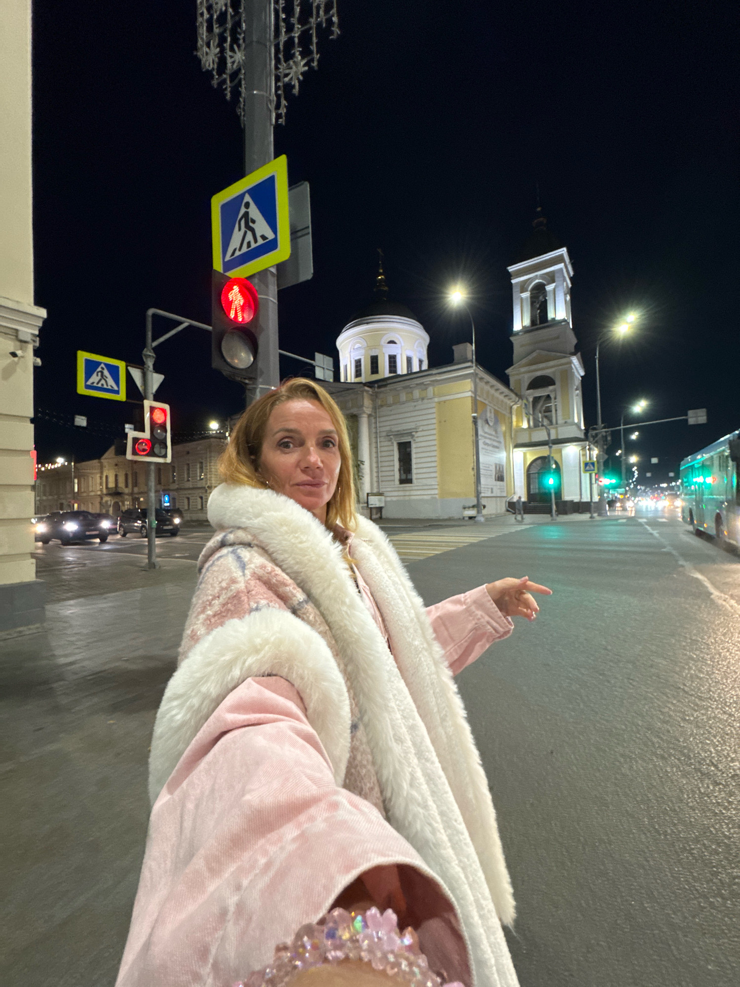 A blonde woman in a pink coat and white fur-lined collar stands on a city street at night in Tver, Russia. She is at a crosswalk, indicated by a pedestrian crossing sign and a traffic light displaying red for pedestrians. In the background, a yellow church with a tall bell tower is illuminated. Streetlights and car headlights create a nighttime urban atmosphere. The woman is holding a phone and appears to be taking a selfie, with her left hand extended towards the camera. Cars are visible on the street, and a bus is approaching in the distance. The ground is wet, suggesting recent rain.
