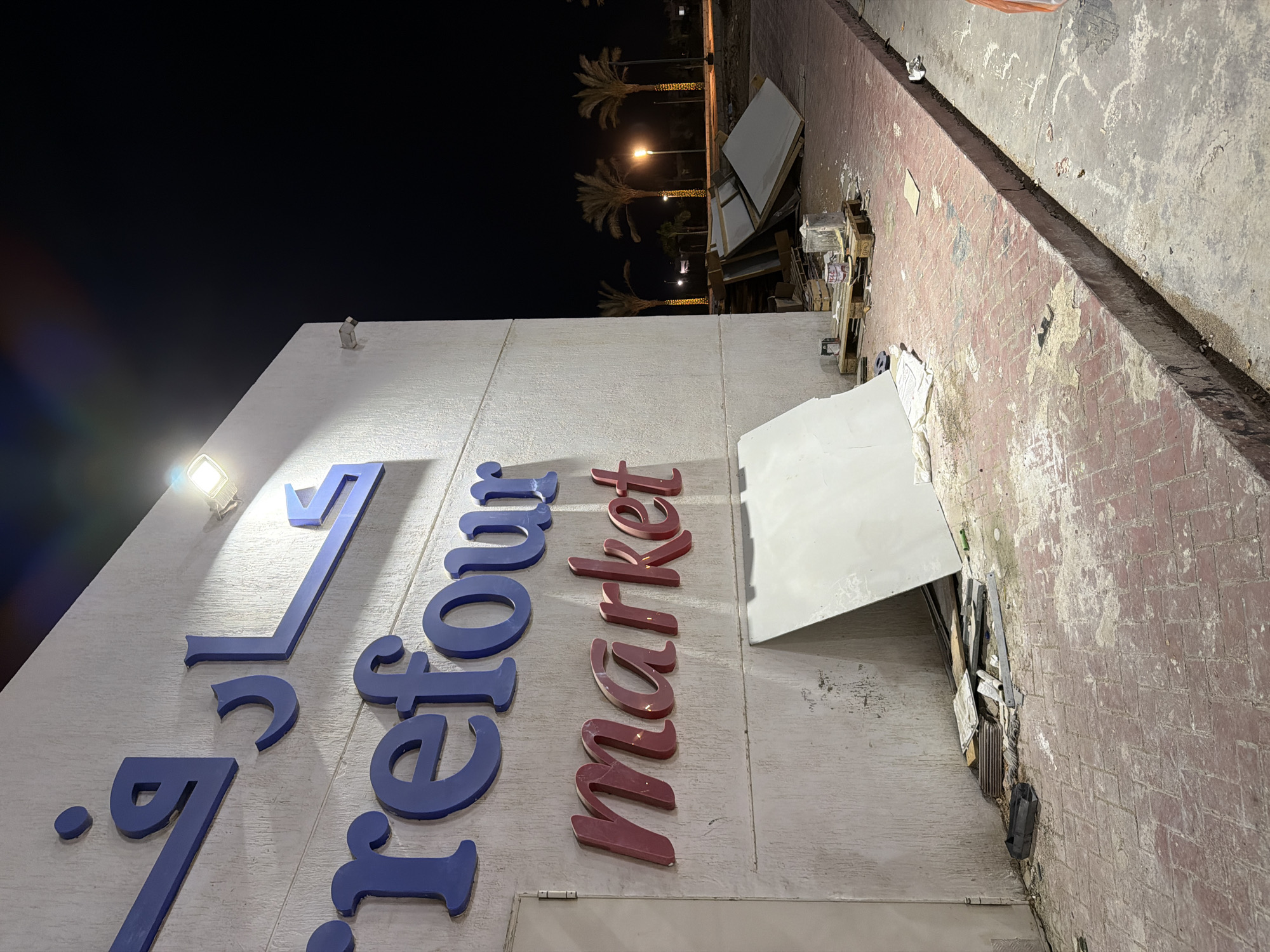 The image shows the exterior of a Carrefour Market store at night in Sharm El Sheikh, Egypt. The store has large blue Arabic script letters above the English words "Carrefour market" in red. Construction materials, including large white panels and wooden pallets, are stacked on the ground in front of the store. The area is paved with red bricks, and there is debris scattered on the ground. Palm trees are visible in the background, and streetlights are on. The scene appears to be a construction or renovation site, with some signs of disarray.